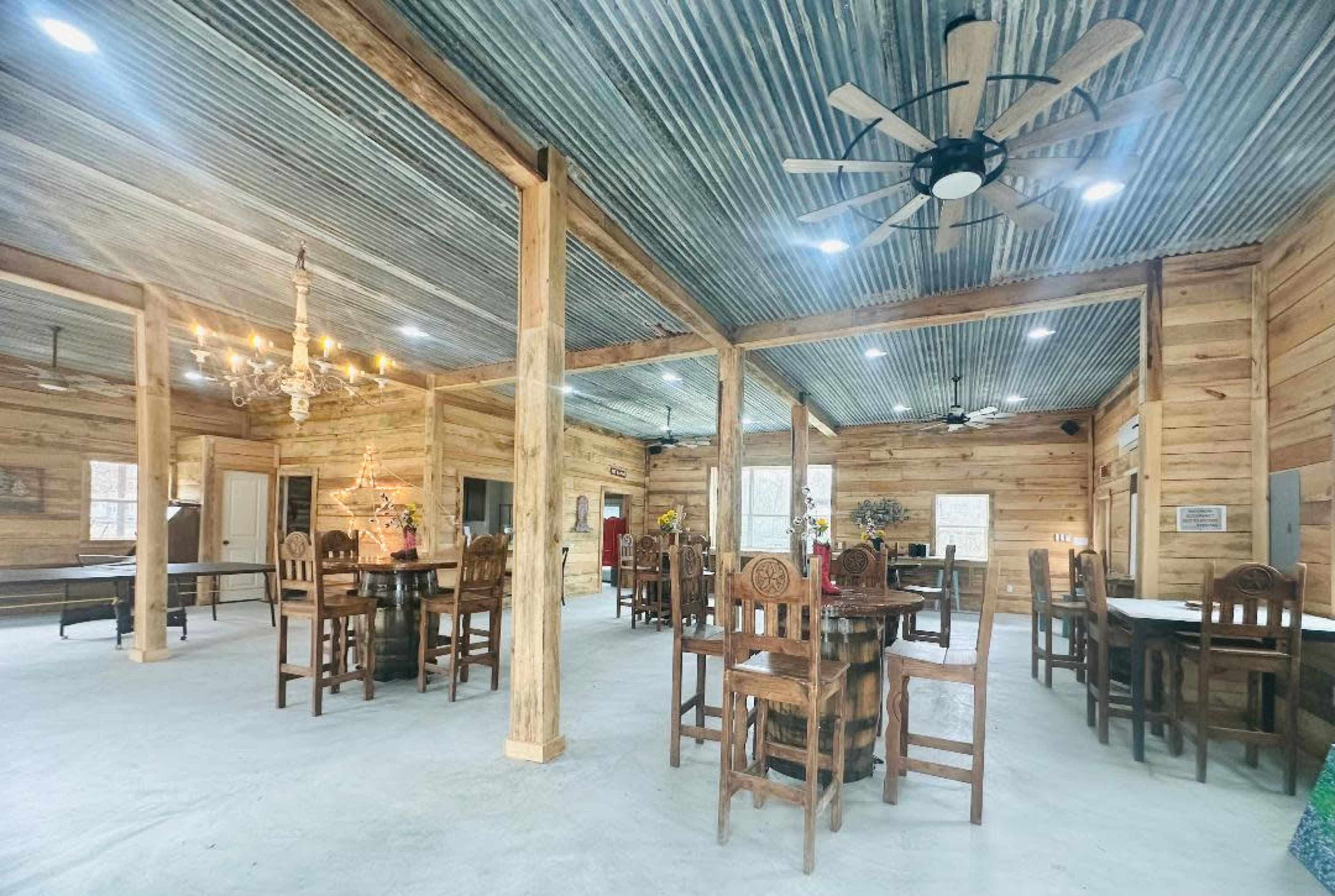 A spacious wooden interior with metal roofing, featuring several dining tables surrounded by chairs and a chandelier hanging from the ceiling.