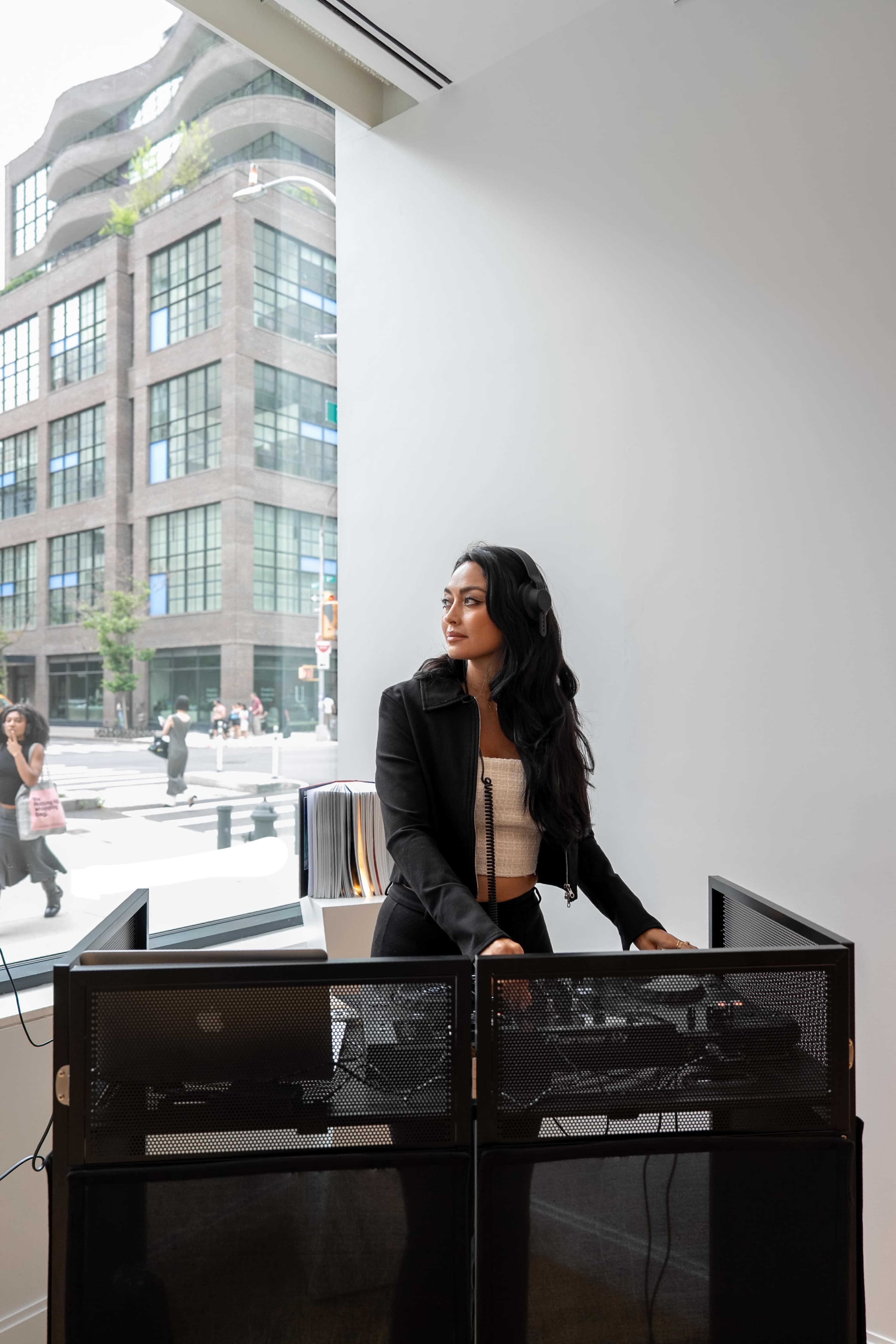 A woman stands at a DJ booth, looking out a large window towards a busy street with pedestrians and buildings in the background.