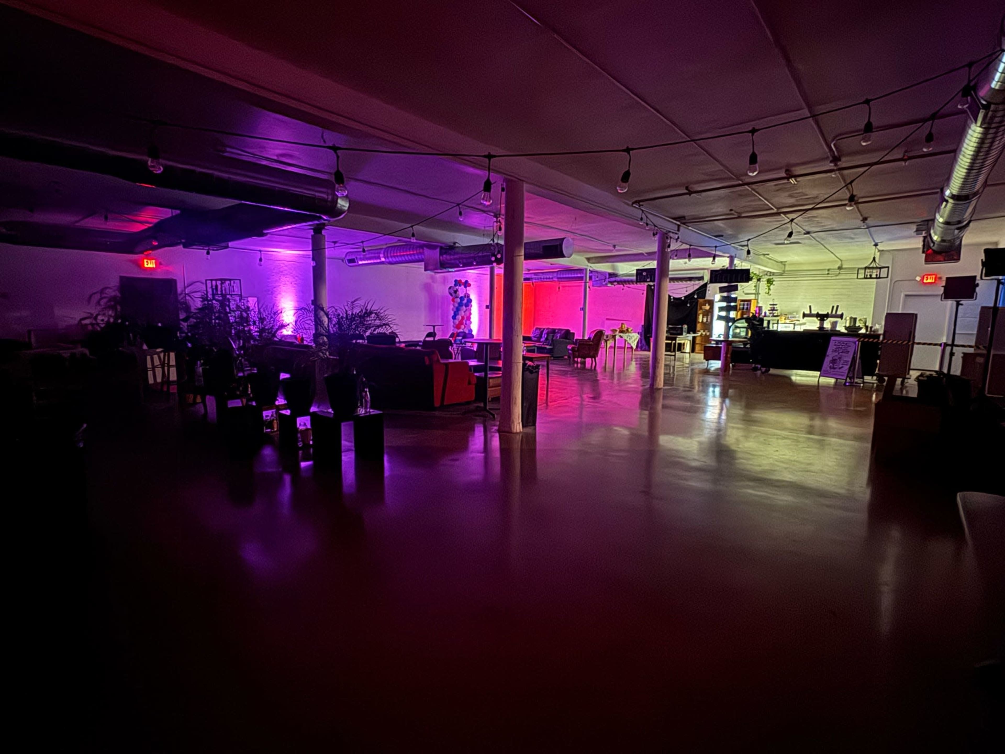 The image shows a dimly lit interior of a versatile space featuring seating areas, plants, and a bar, illuminated by purple and pink lights.