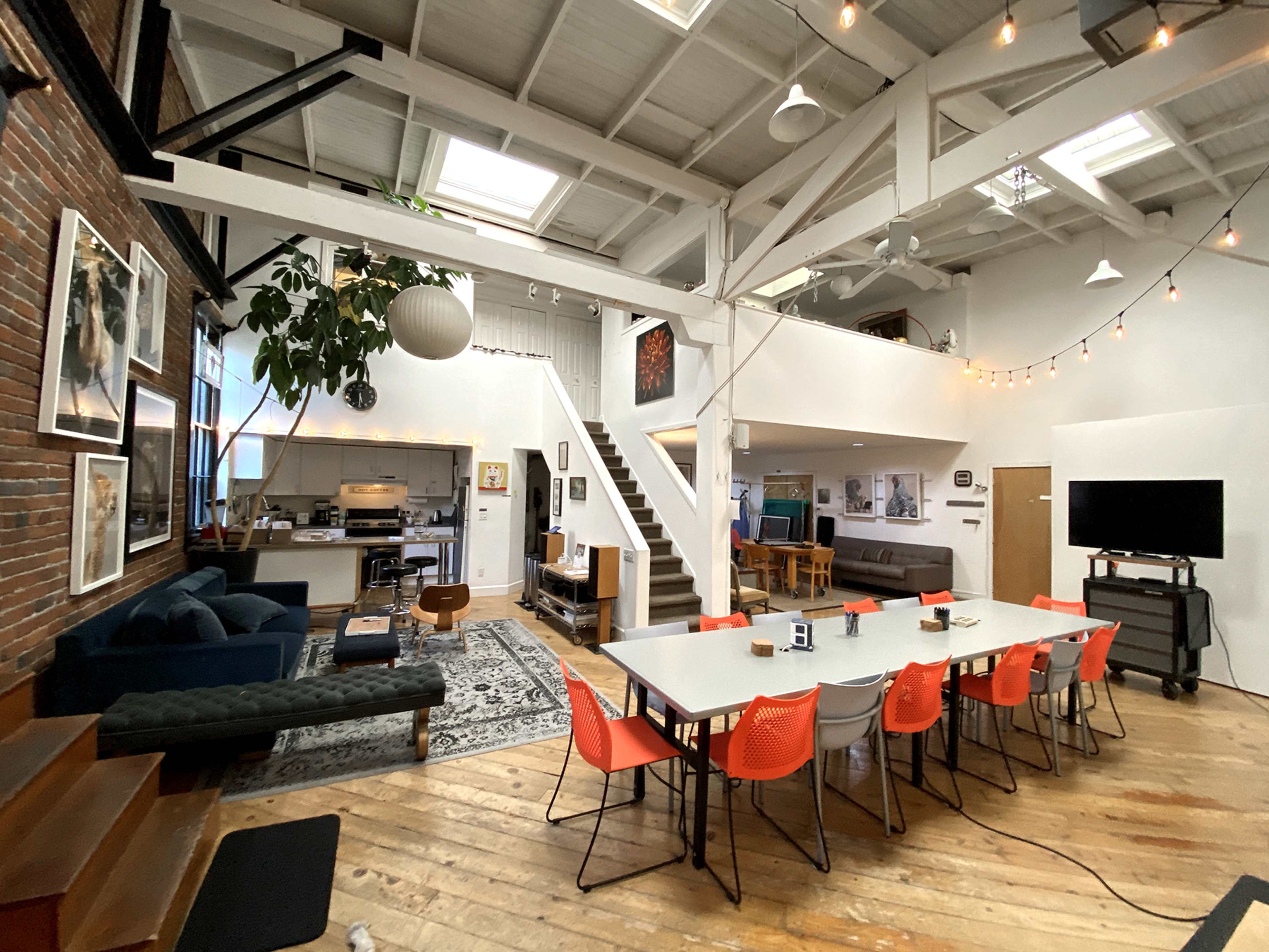 The image shows a spacious, open-plan living area with a dining table surrounded by orange chairs, a kitchen in the background, and a lofted space above.