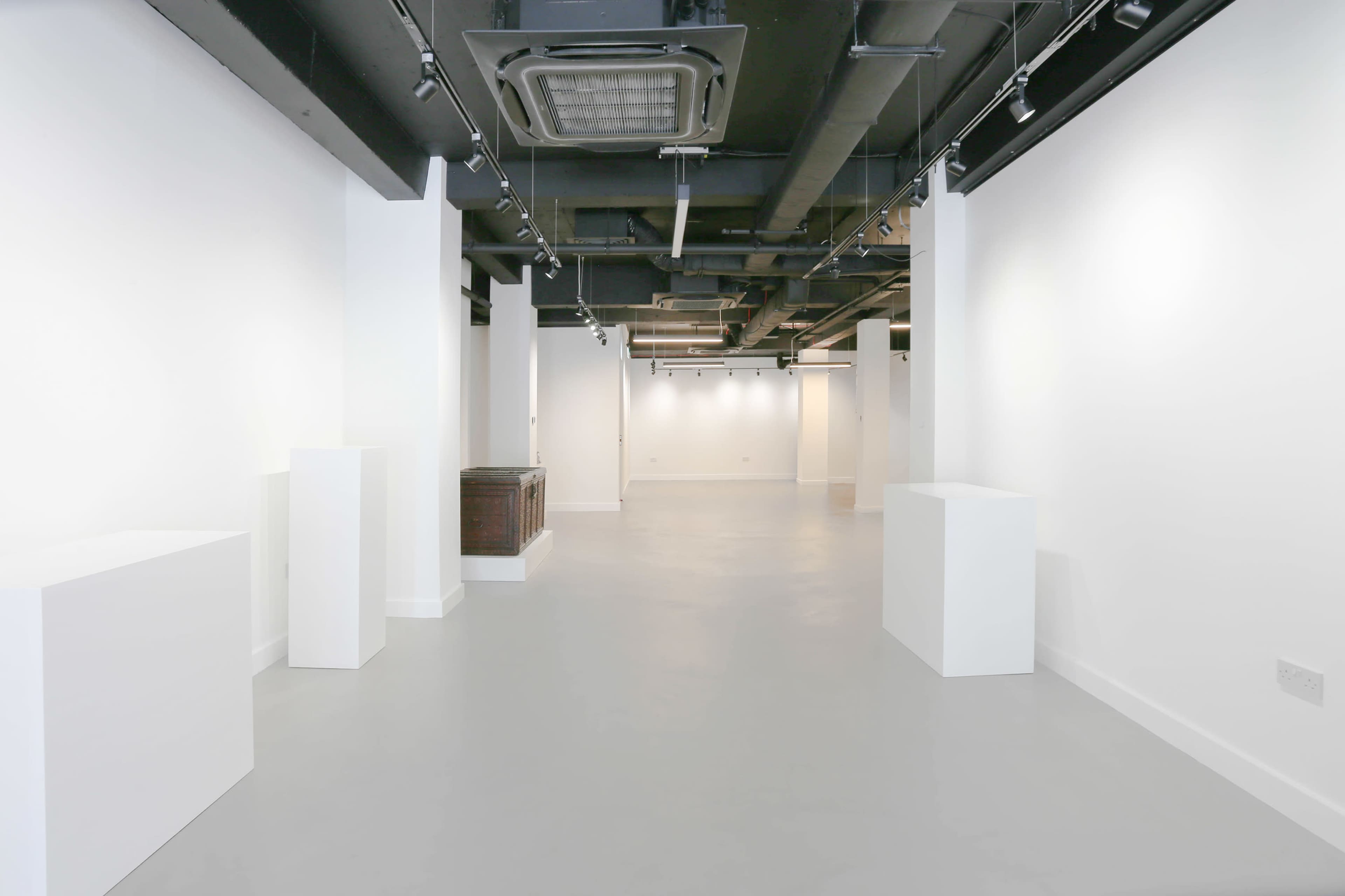 The image shows a spacious, well-lit gallery or exhibition space with empty white walls and several white pedestals arranged on a gray floor.