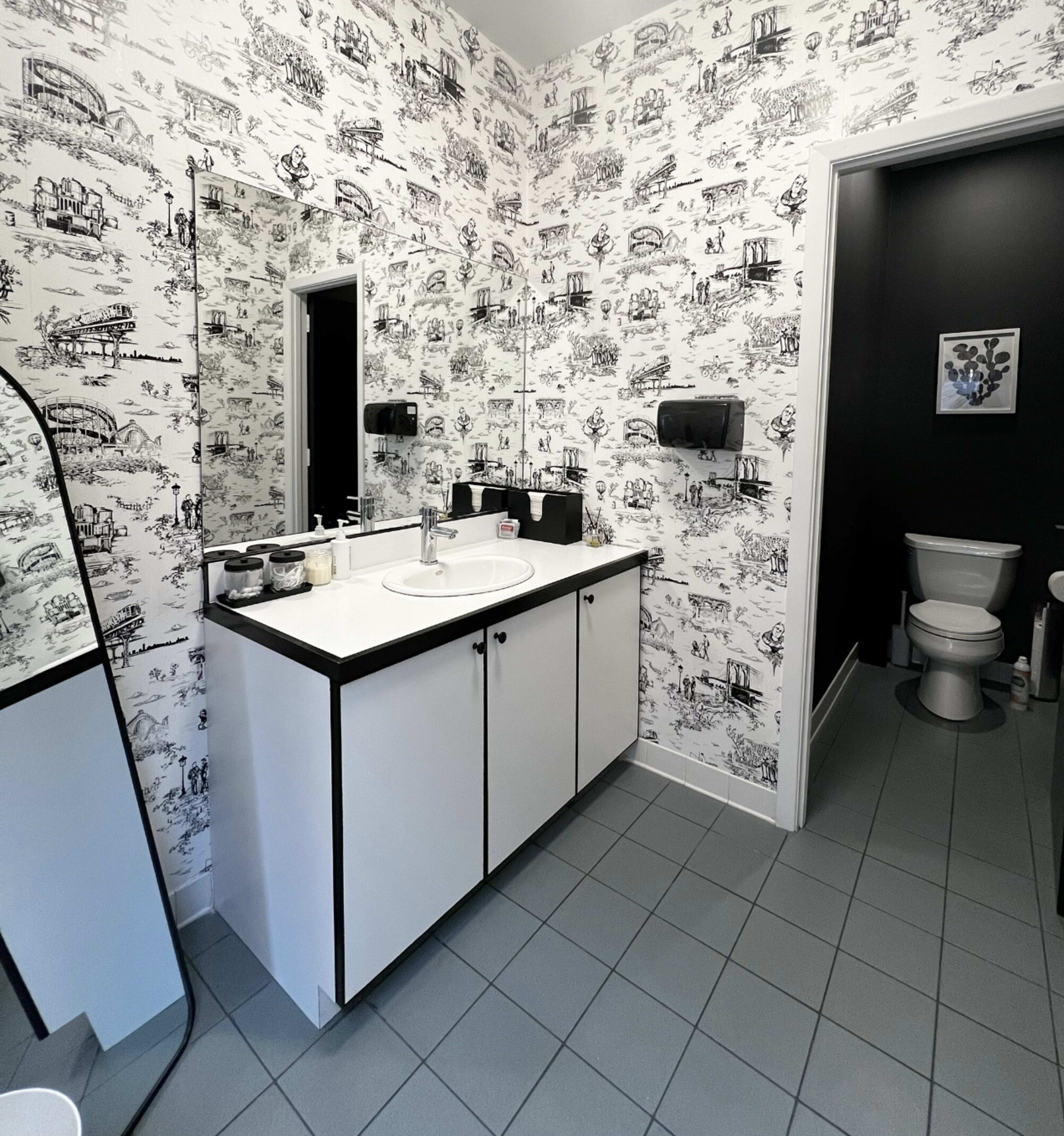 A bathroom with patterned wallpaper, a sink and vanity on one side, and a toilet in a separate room.