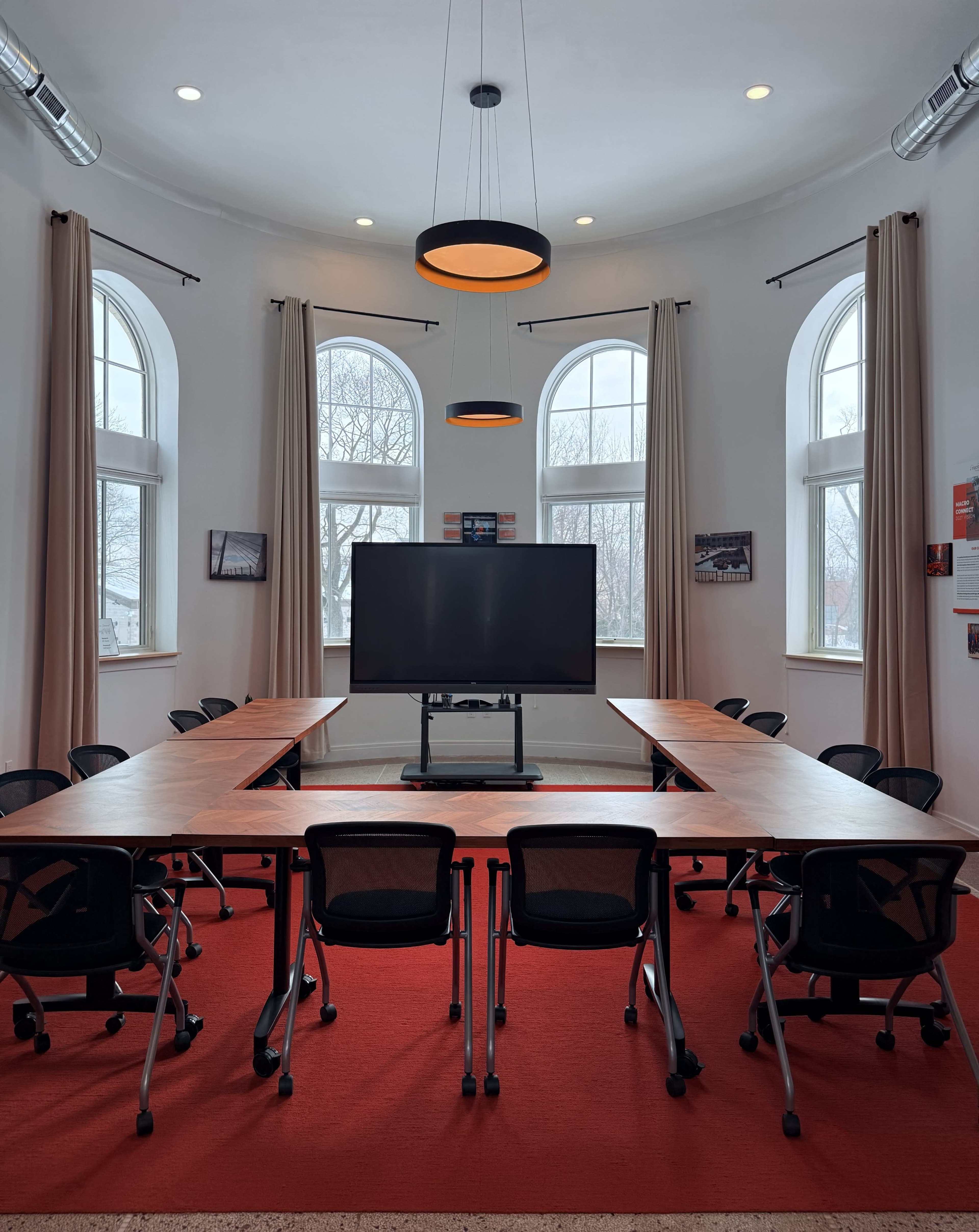 The image shows a modern meeting room with a large screen at the front, surrounded by a U-shaped arrangement of wooden tables and black chairs on a red carpet.