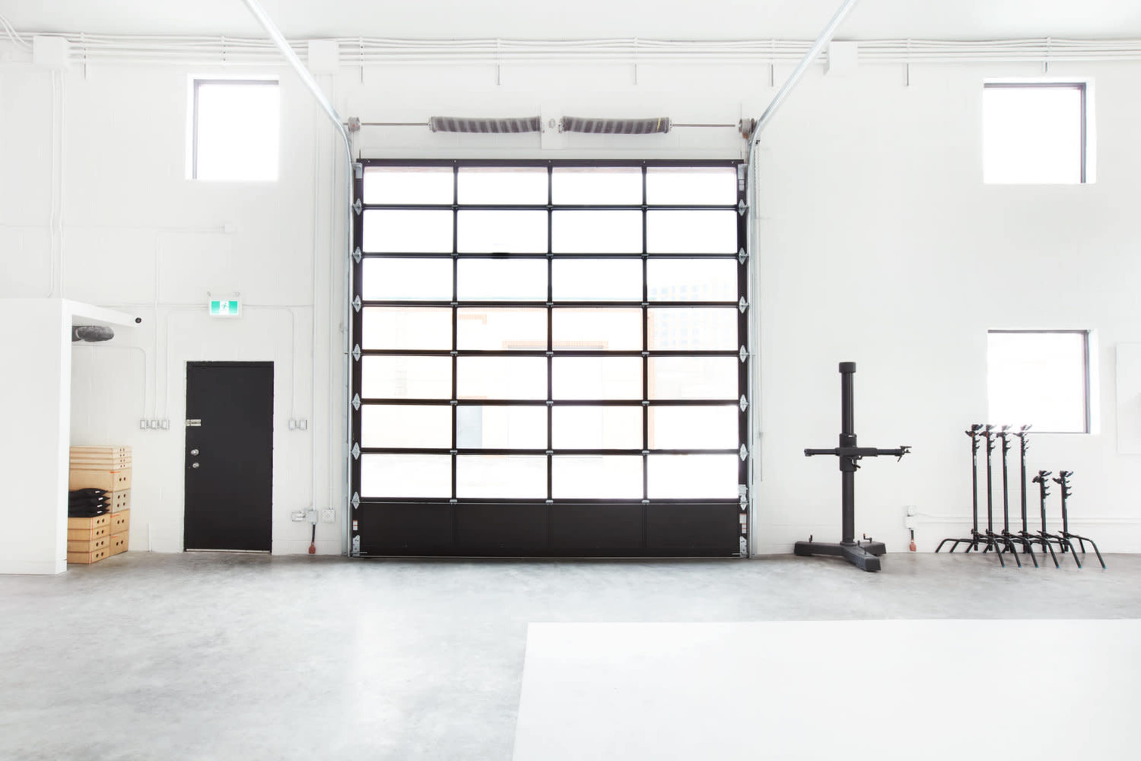 A spacious, minimalist interior with a large garage door, black door, and several exercise equipment racks against a light-colored wall.