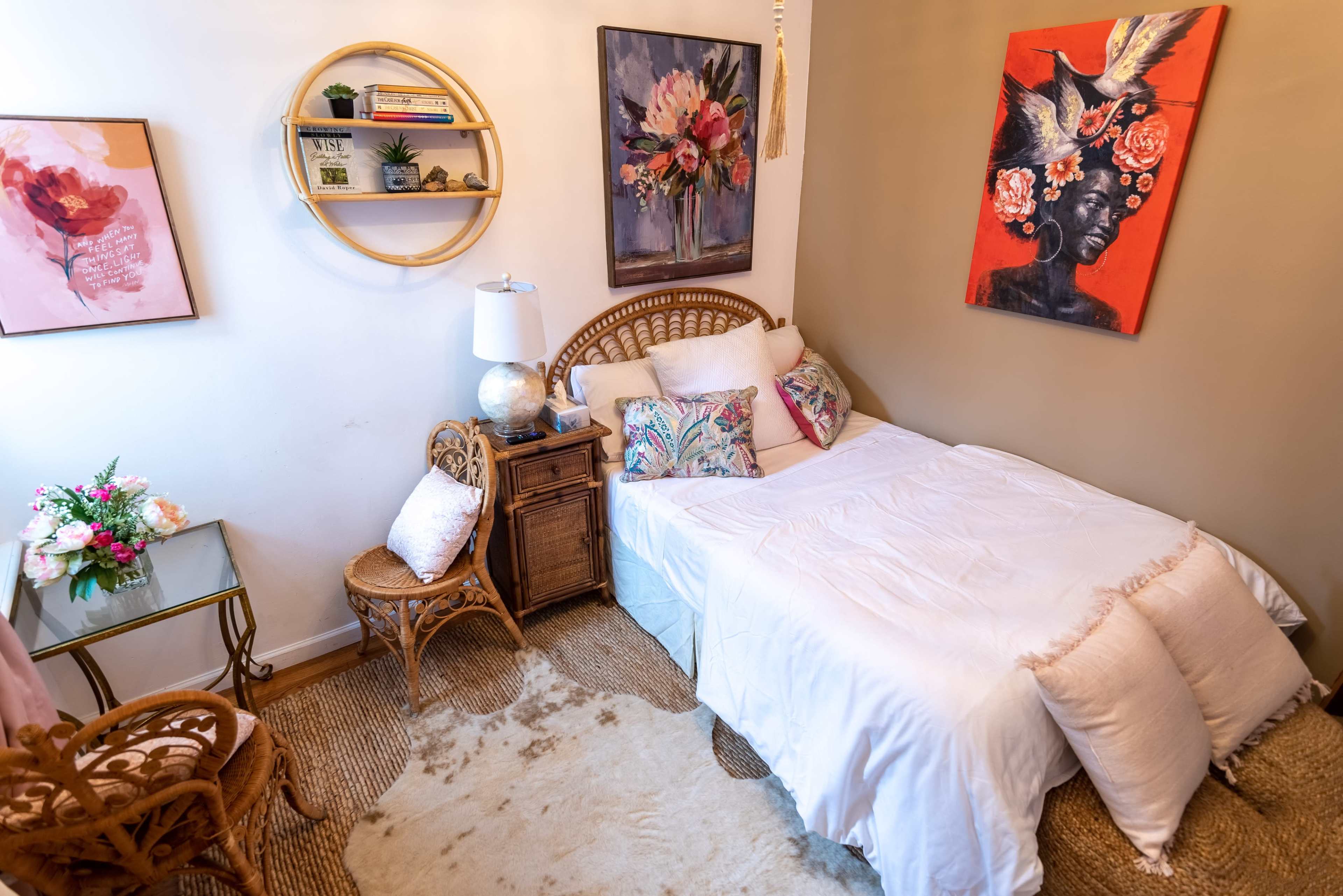 The room features a bed with white bedding, a rattan chair, a small glass-topped table, and colorful artwork on the walls.