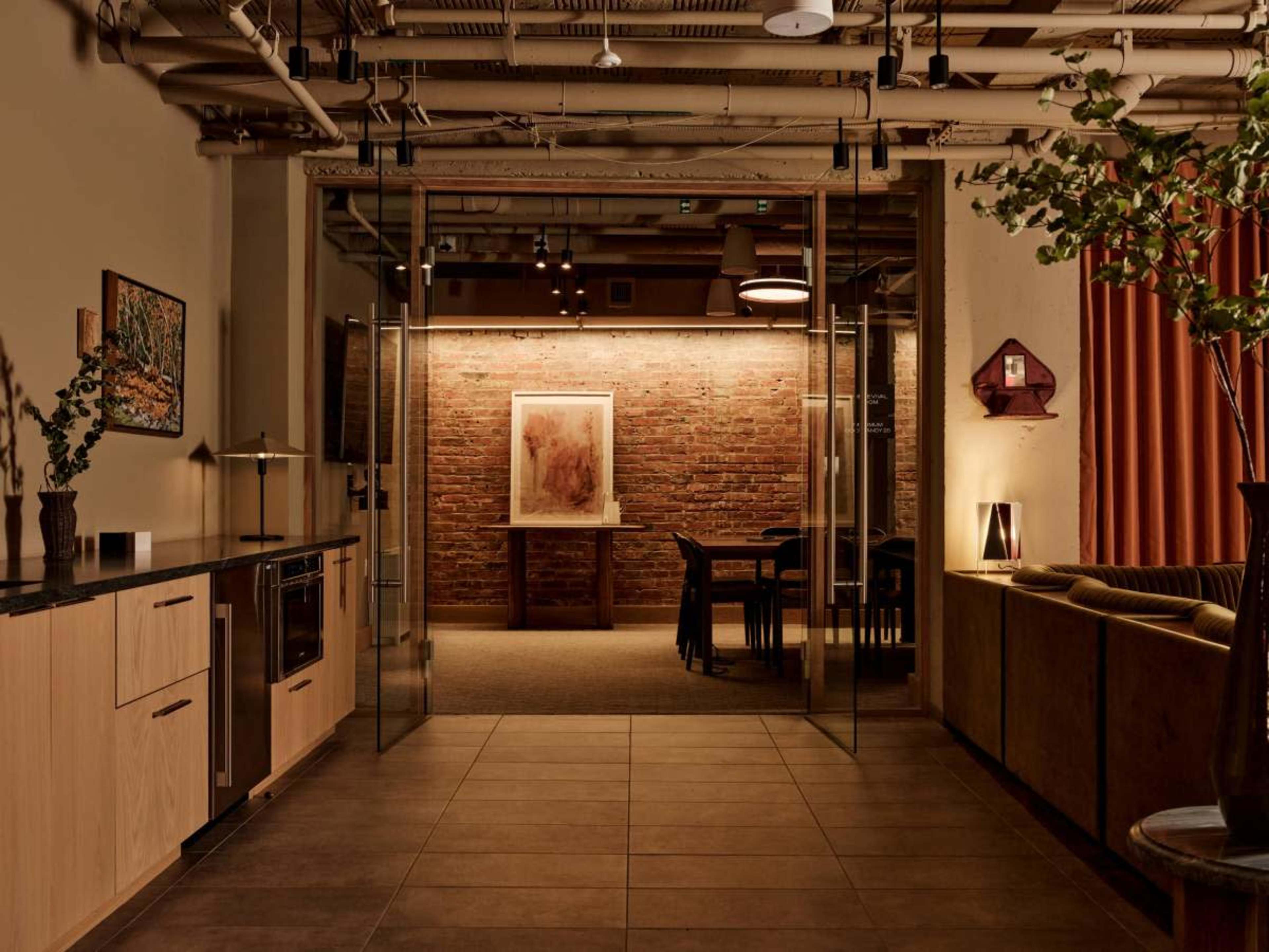 The image shows a contemporary interior space with a kitchen area on the left, a glass partition leading to a dining area with a brick wall, and a seating area on the right.