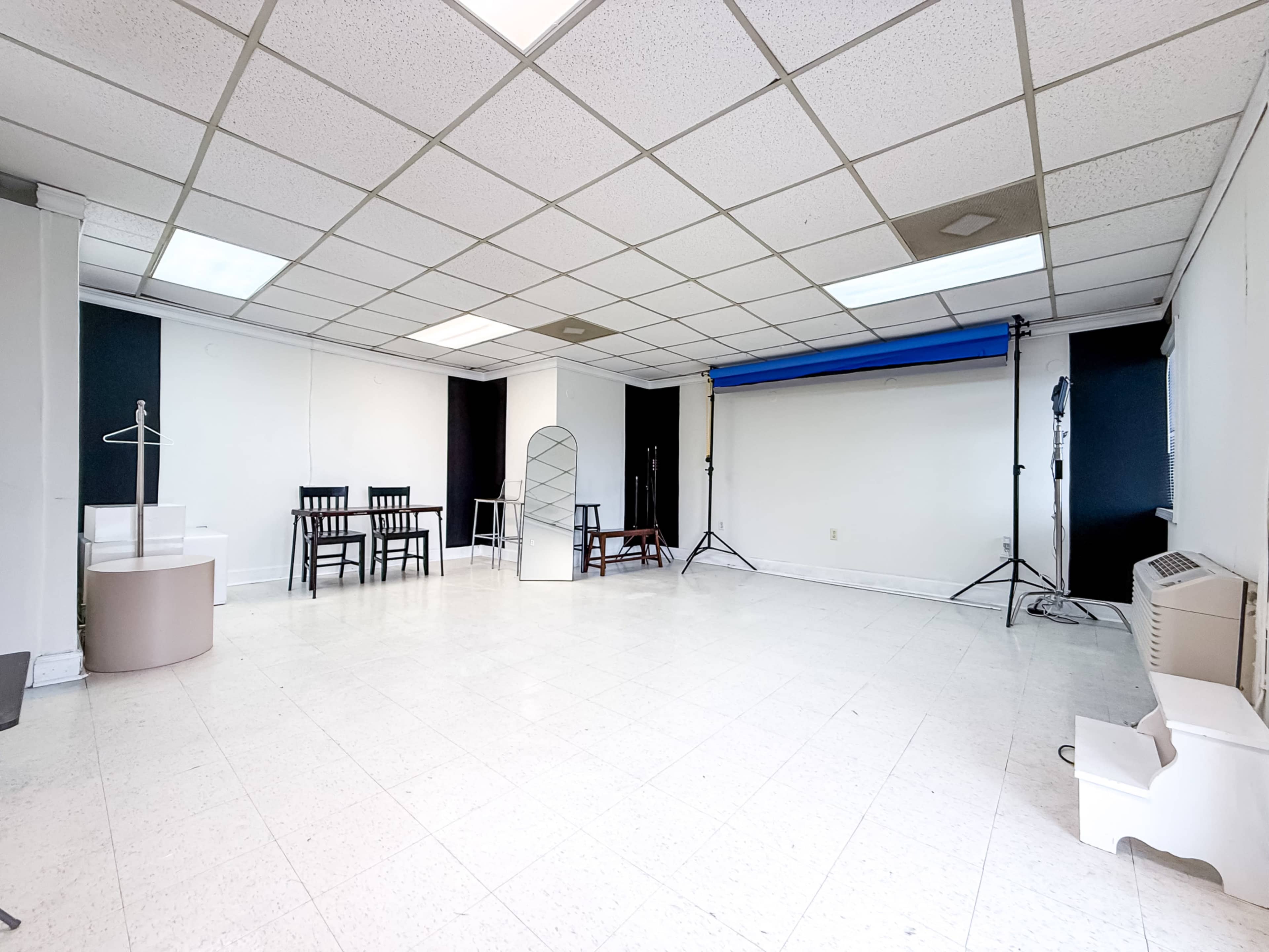 The image shows an empty studio space with white walls, a tiled floor, and several furniture pieces, including chairs and a mirror, arranged along one side.