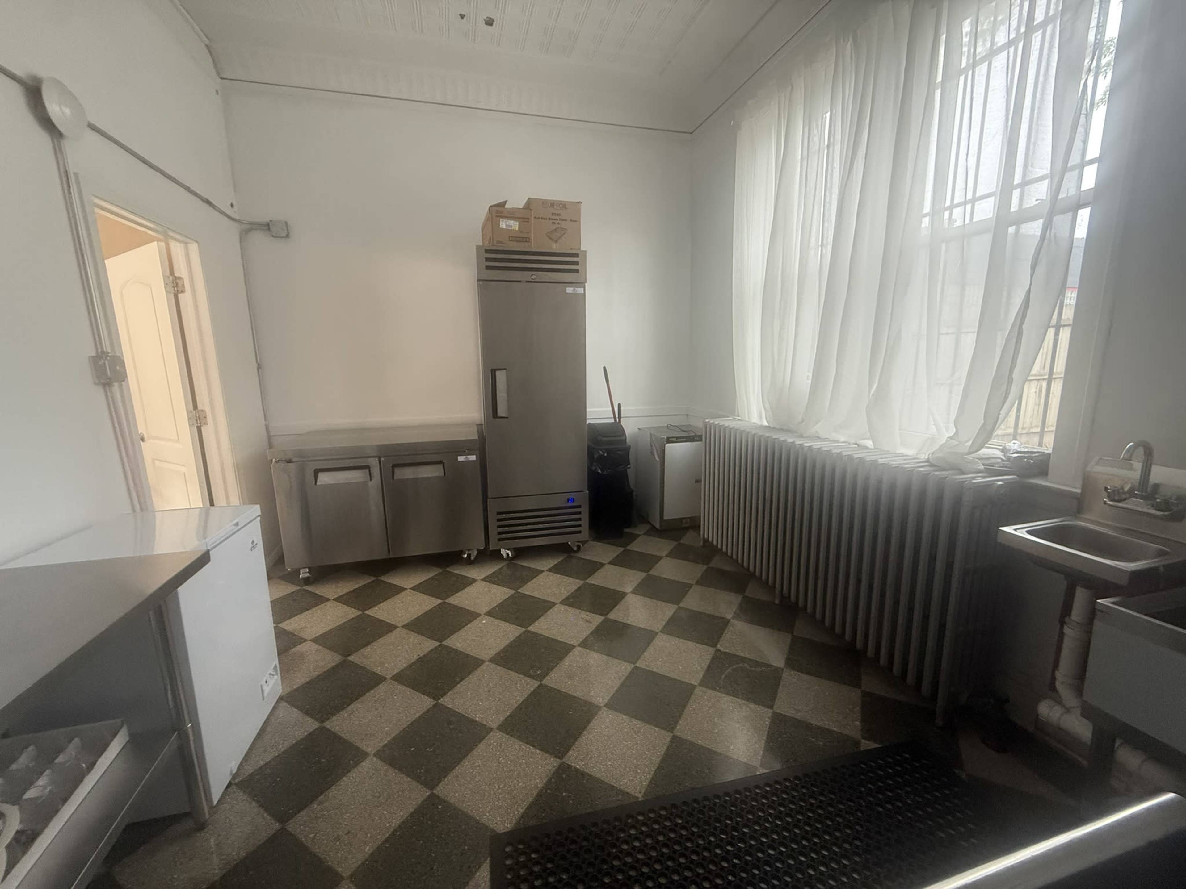 The image shows a kitchen area with a refrigerator, several ice bins, a sink, and a large window letting in light, all set against a checkered floor.