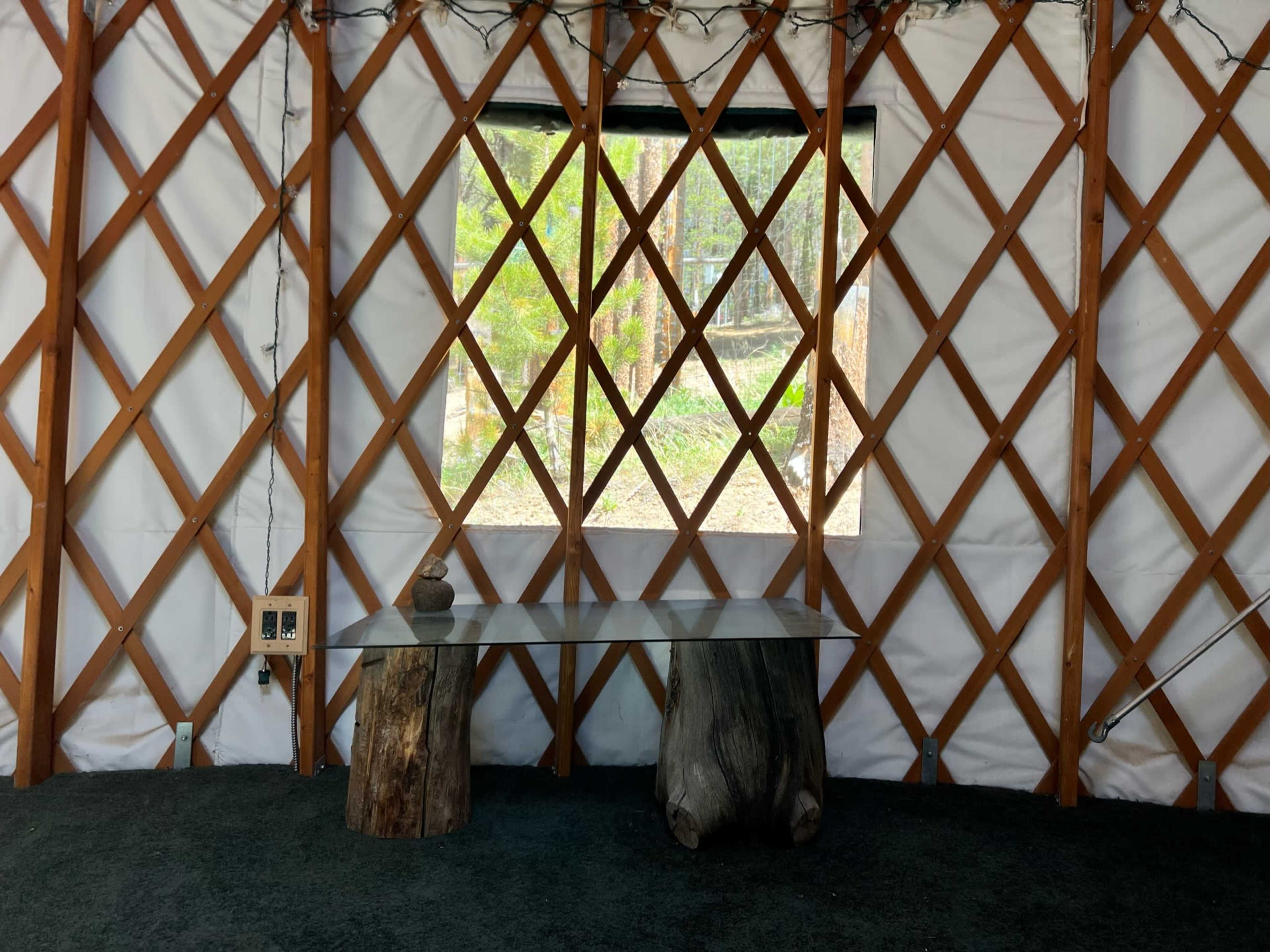 The interior of a yurt features two wooden stools and a glass table beneath a large window framed by lattice wooden beams.