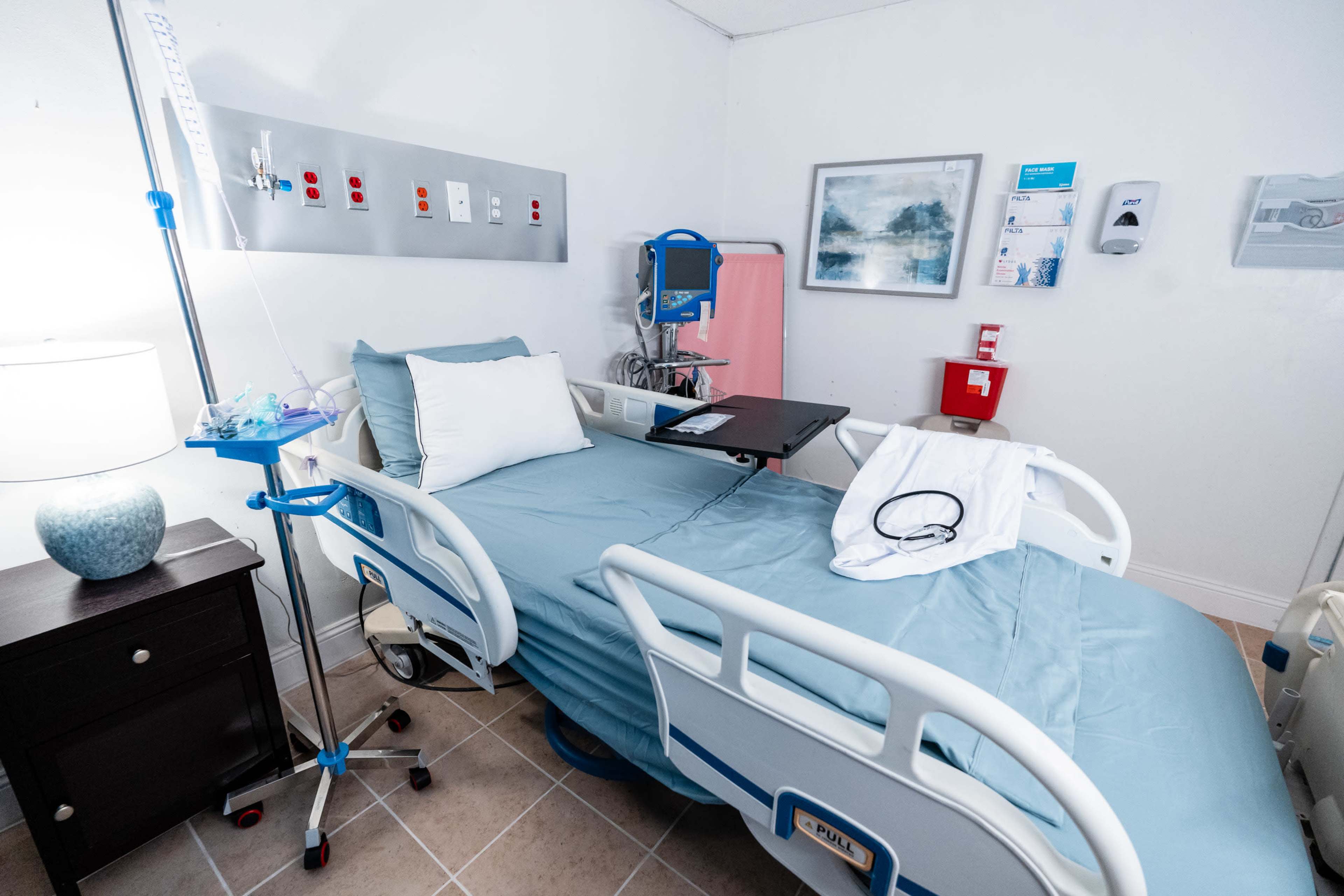 The image shows a hospital room featuring a patient bed with light blue linens, medical equipment, and a small bedside table with a lamp.