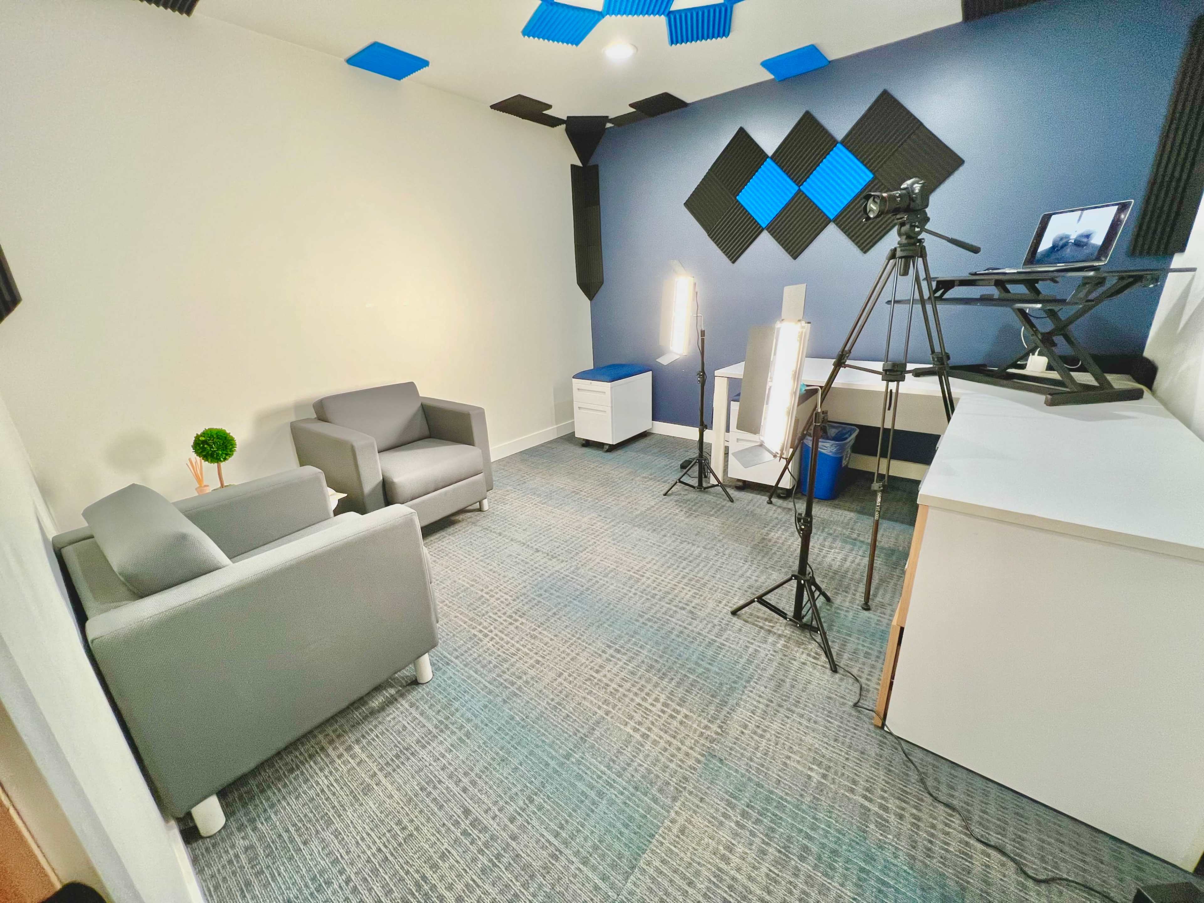 A small studio setup with two gray chairs, a white desk, a camera on a tripod, and lighting equipment against a blue and white wall.