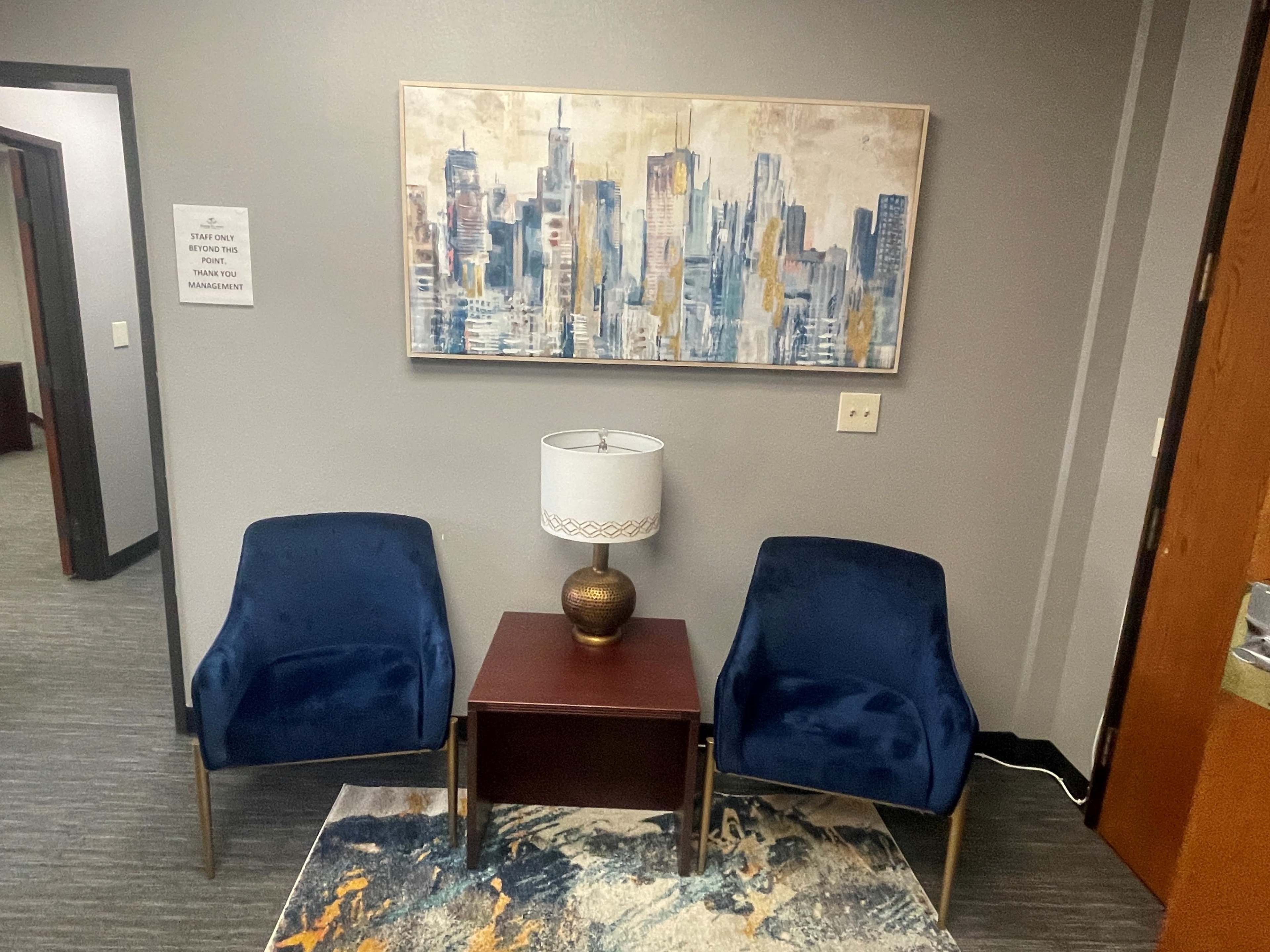 A small waiting area with two blue chairs, a lamp on a wooden table, and a cityscape painting on the wall.