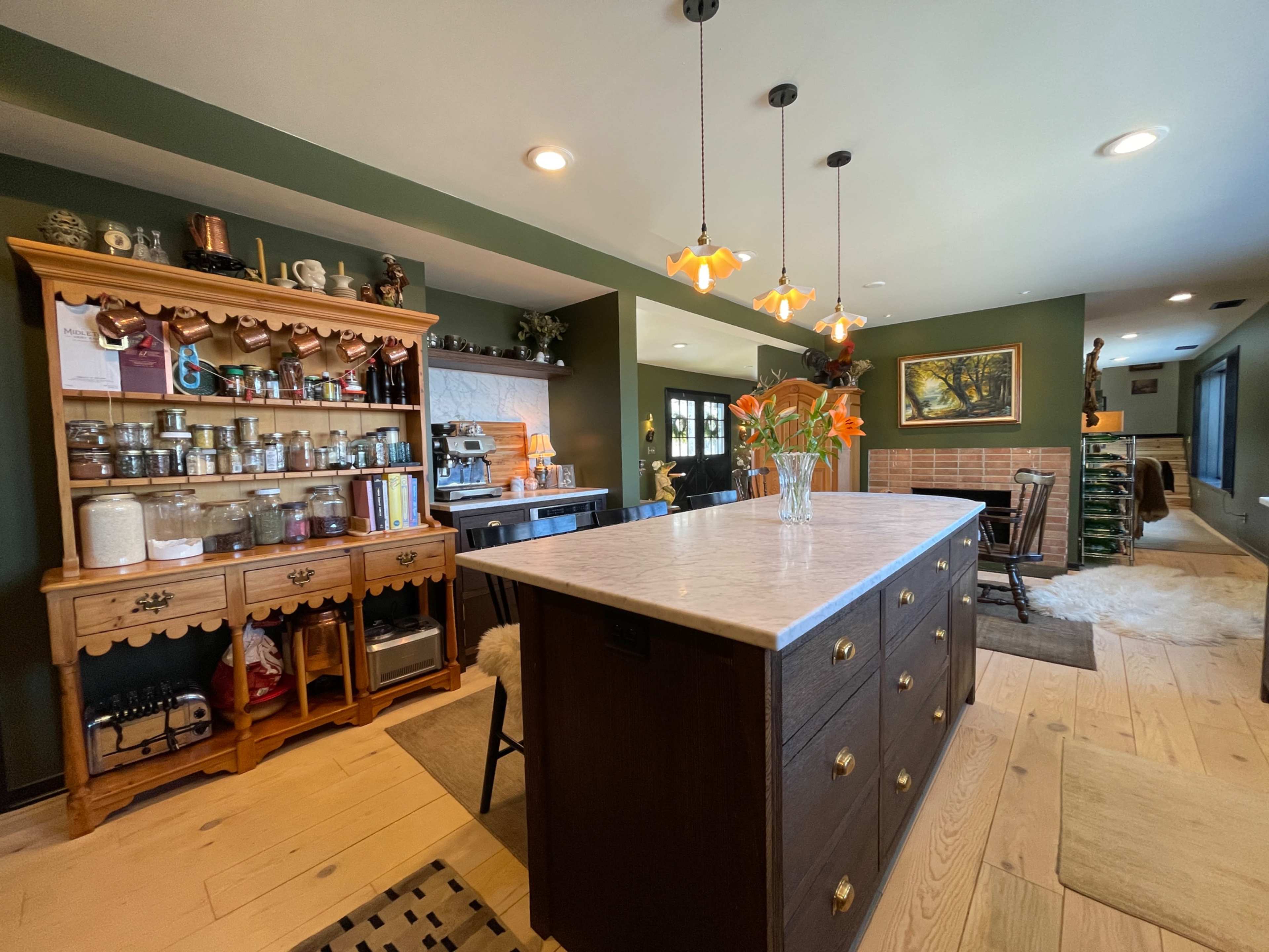 The kitchen features a central island with a light-colored countertop, surrounded by wooden cabinetry and a glass-fronted display for jars and cooking supplies.