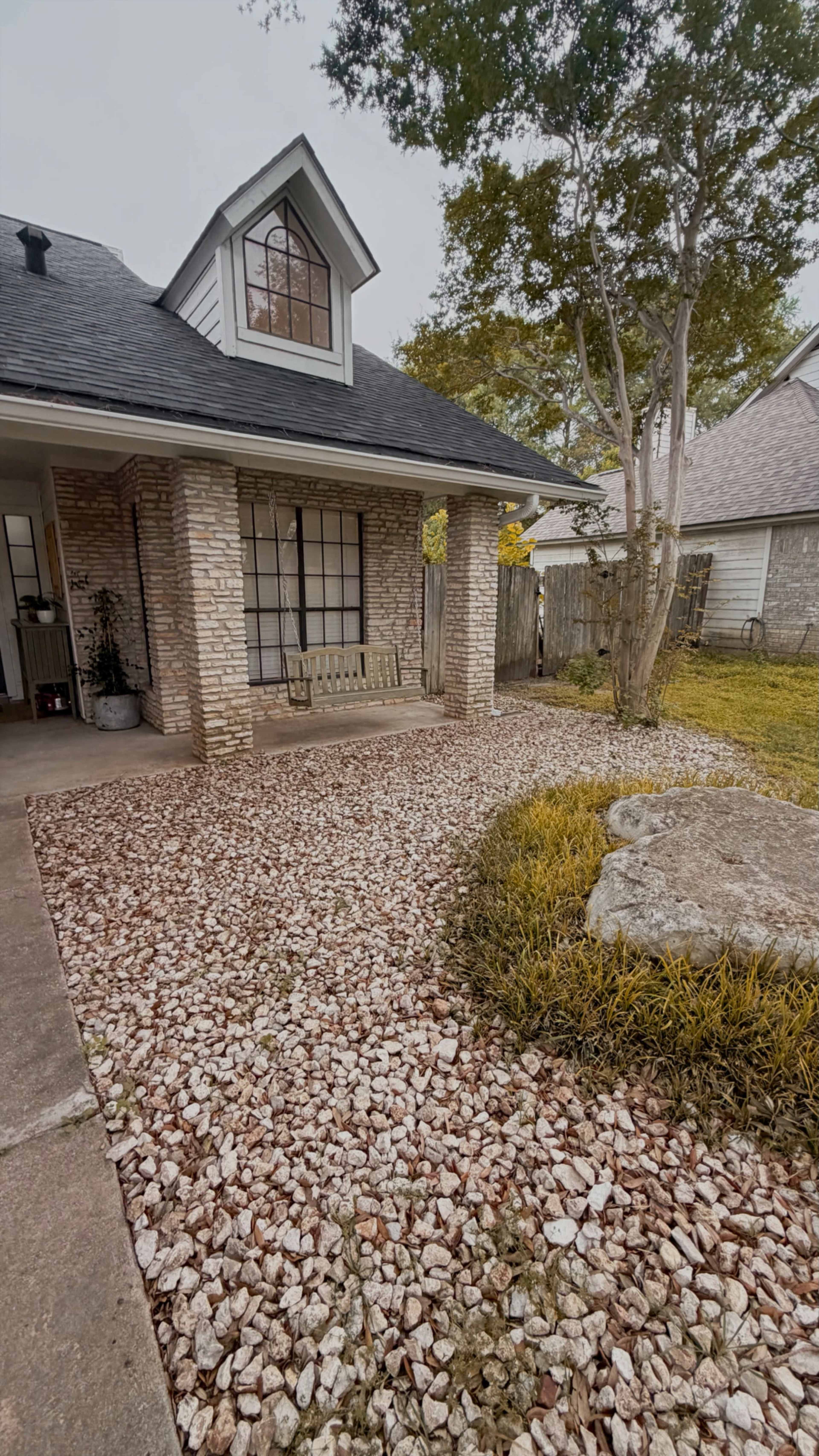 The image shows a house with a stone façade, featuring a front porch with a bench and a landscaped yard containing decorative rocks and grass.