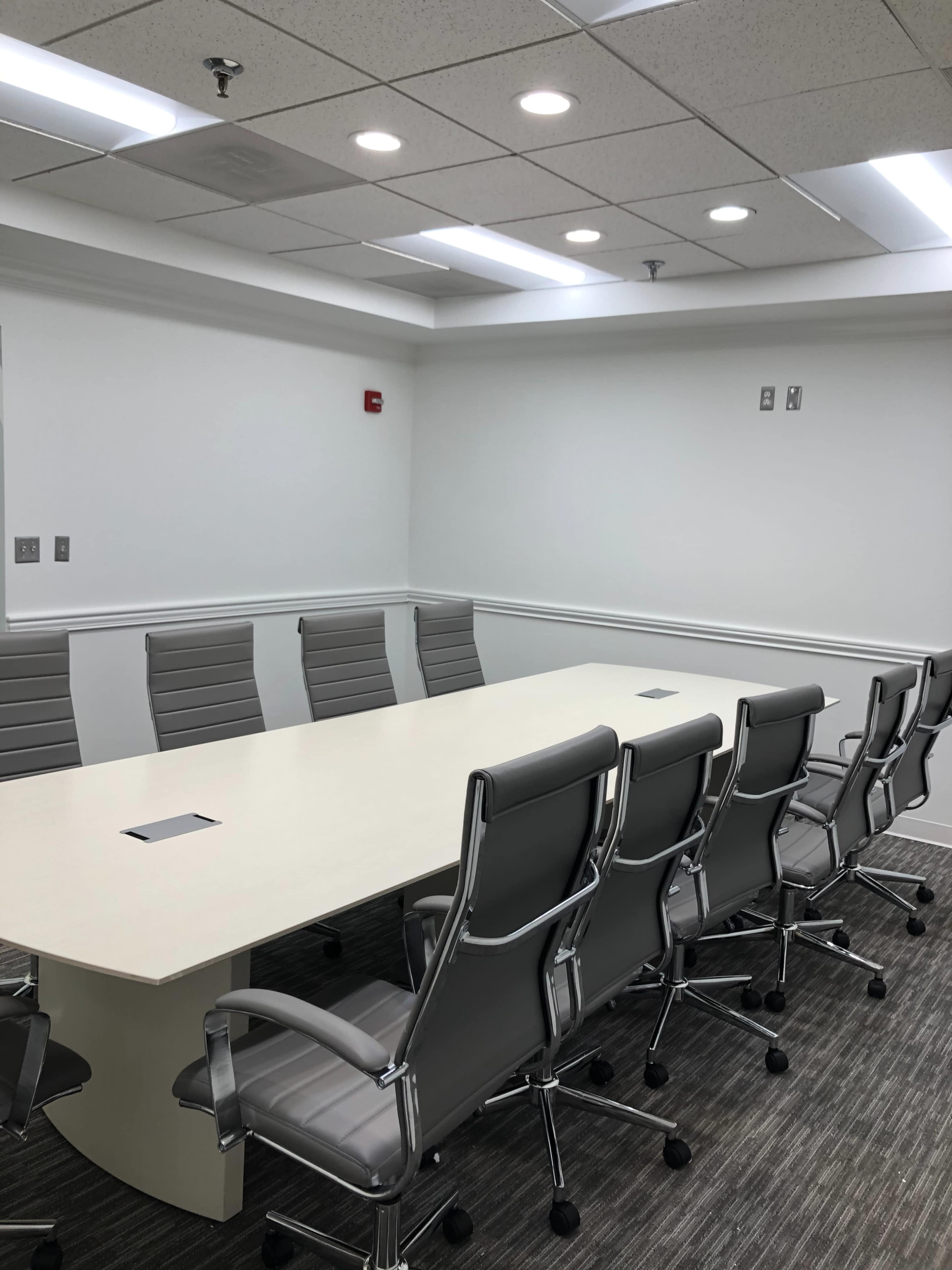 A modern conference room features a long table surrounded by ergonomic chairs, with overhead lighting and wall-mounted outlets.