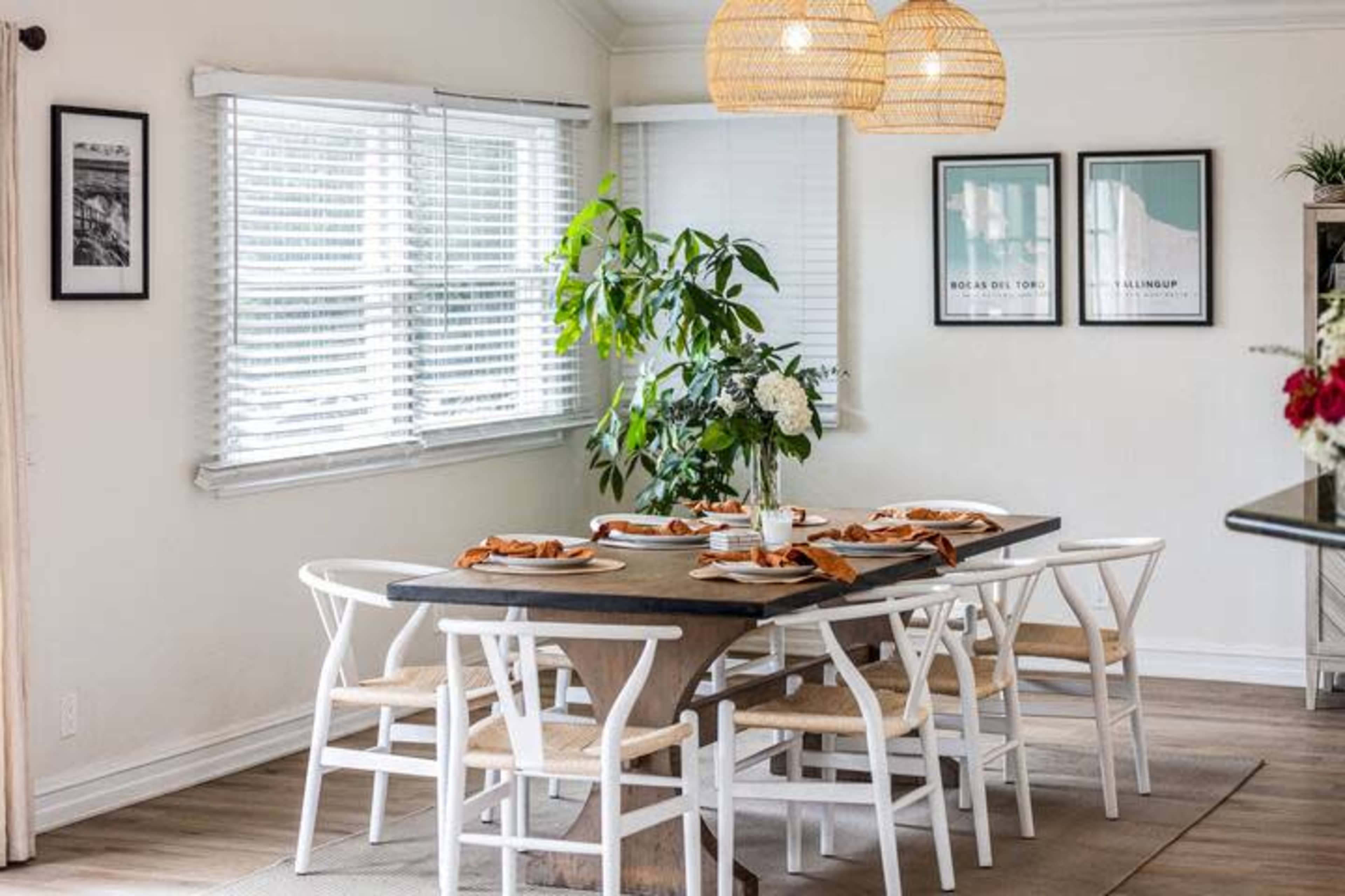 A modern dining room features a large wooden table set for a meal, surrounded by white chairs and illuminated by pendant lights.
