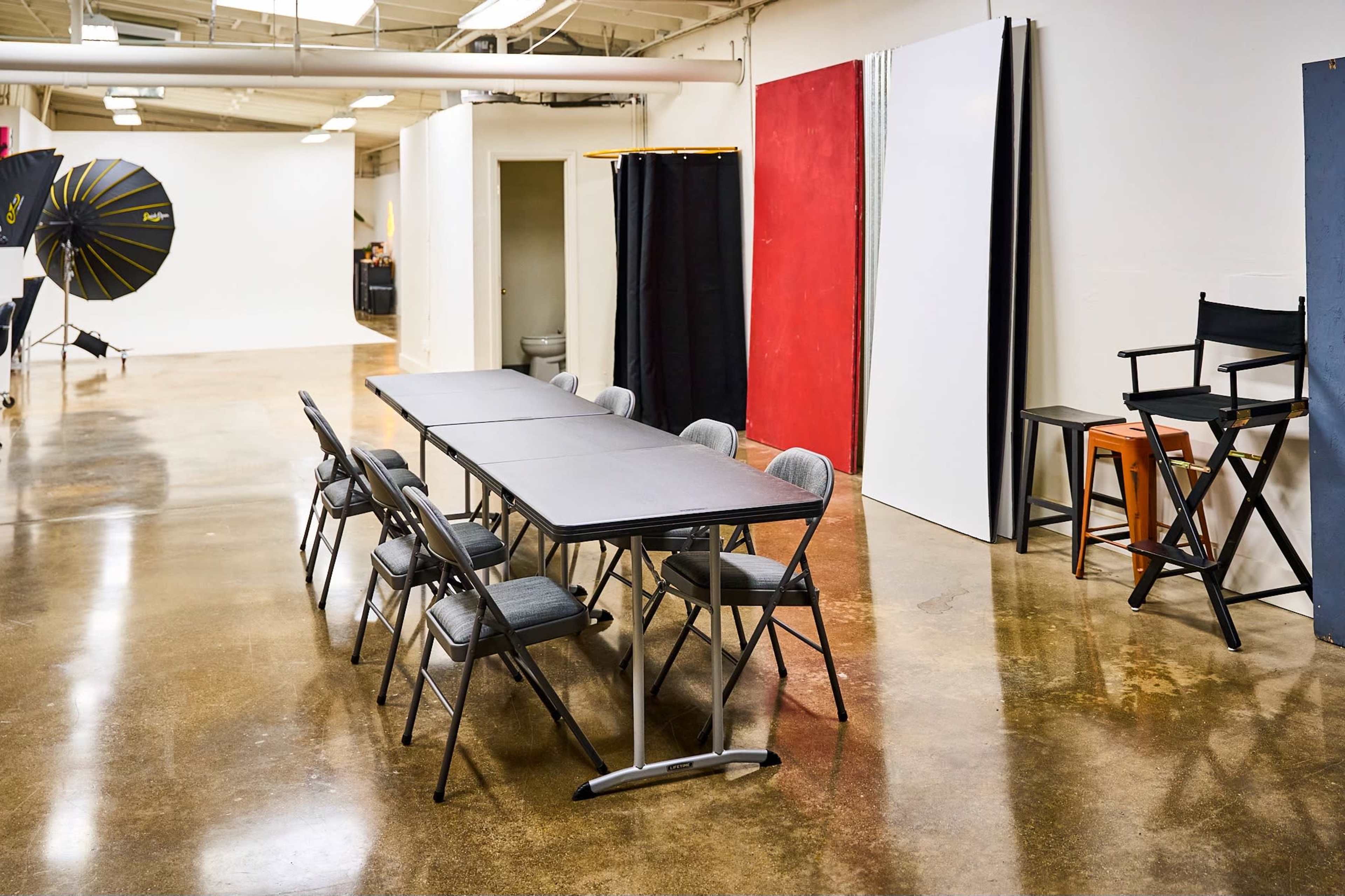A spacious photography studio features a row of folding tables and chairs, with lighting equipment and backdrops positioned along the walls.