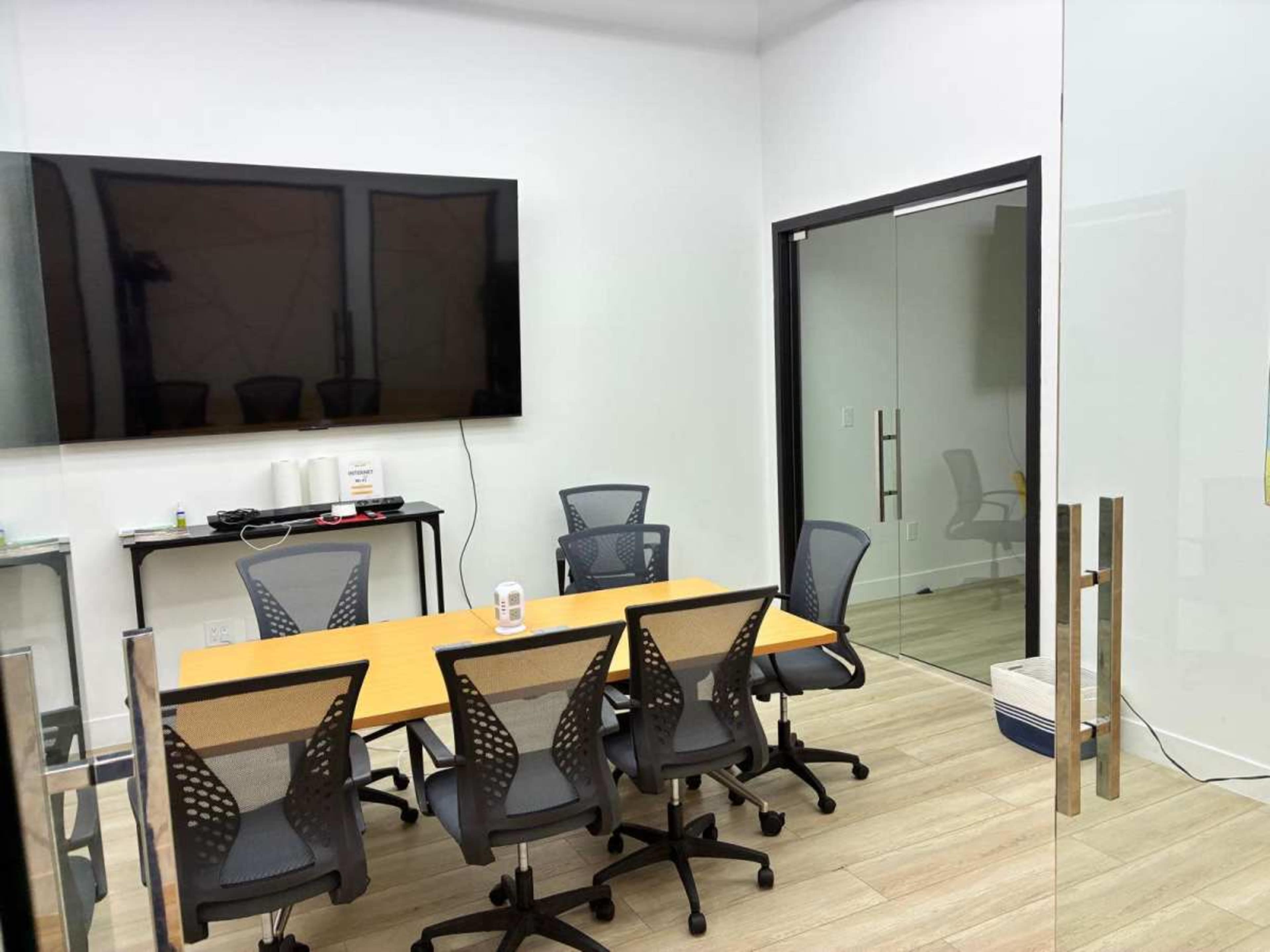 The image shows a modern, empty conference room with a rectangular table surrounded by mesh office chairs and a large television screen mounted on the wall.