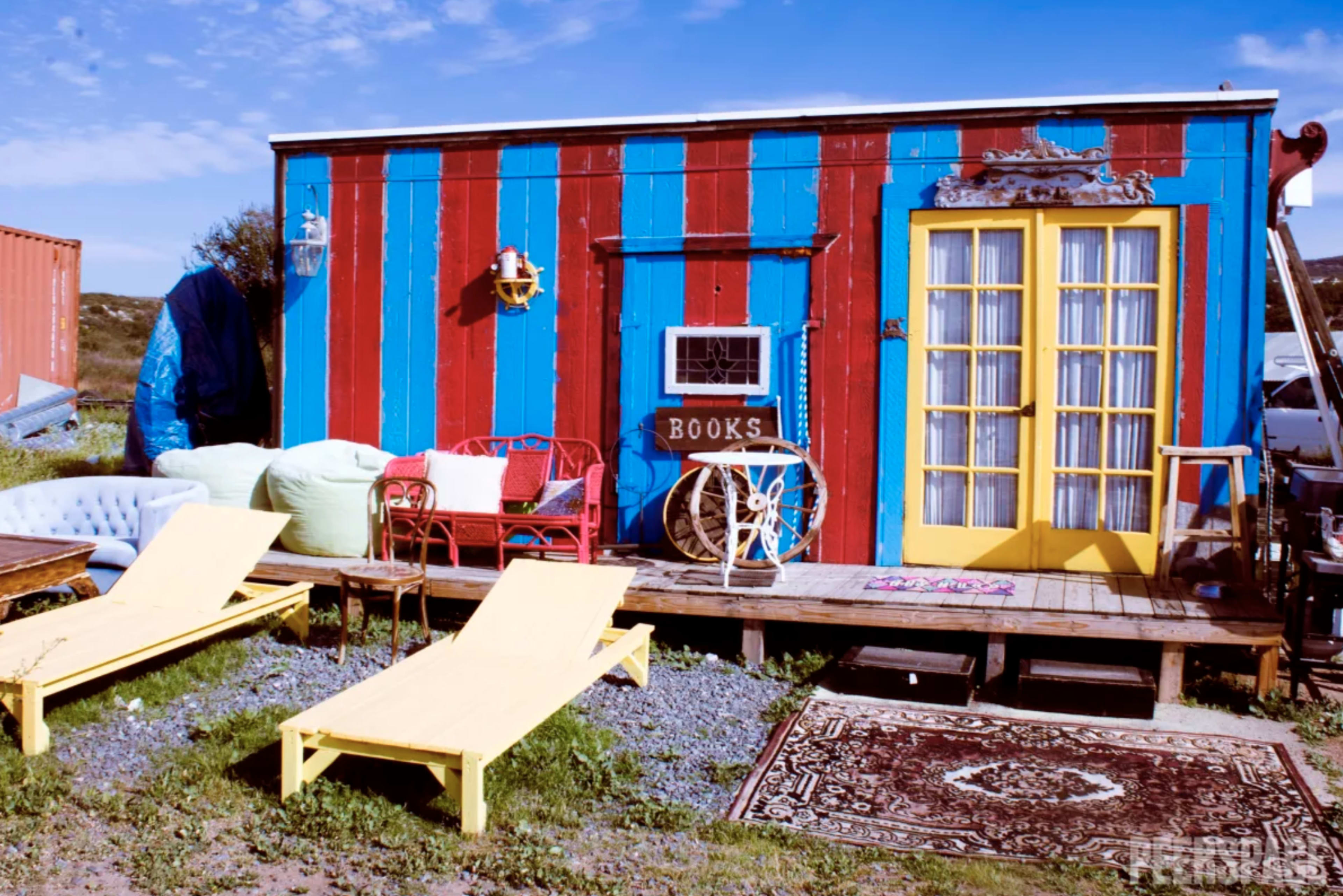 A vibrant, striped cabin with a yellow door and a sign that reads "BOOKS" sits on a wooden deck surrounded by colorful furnishings and outdoor seating.