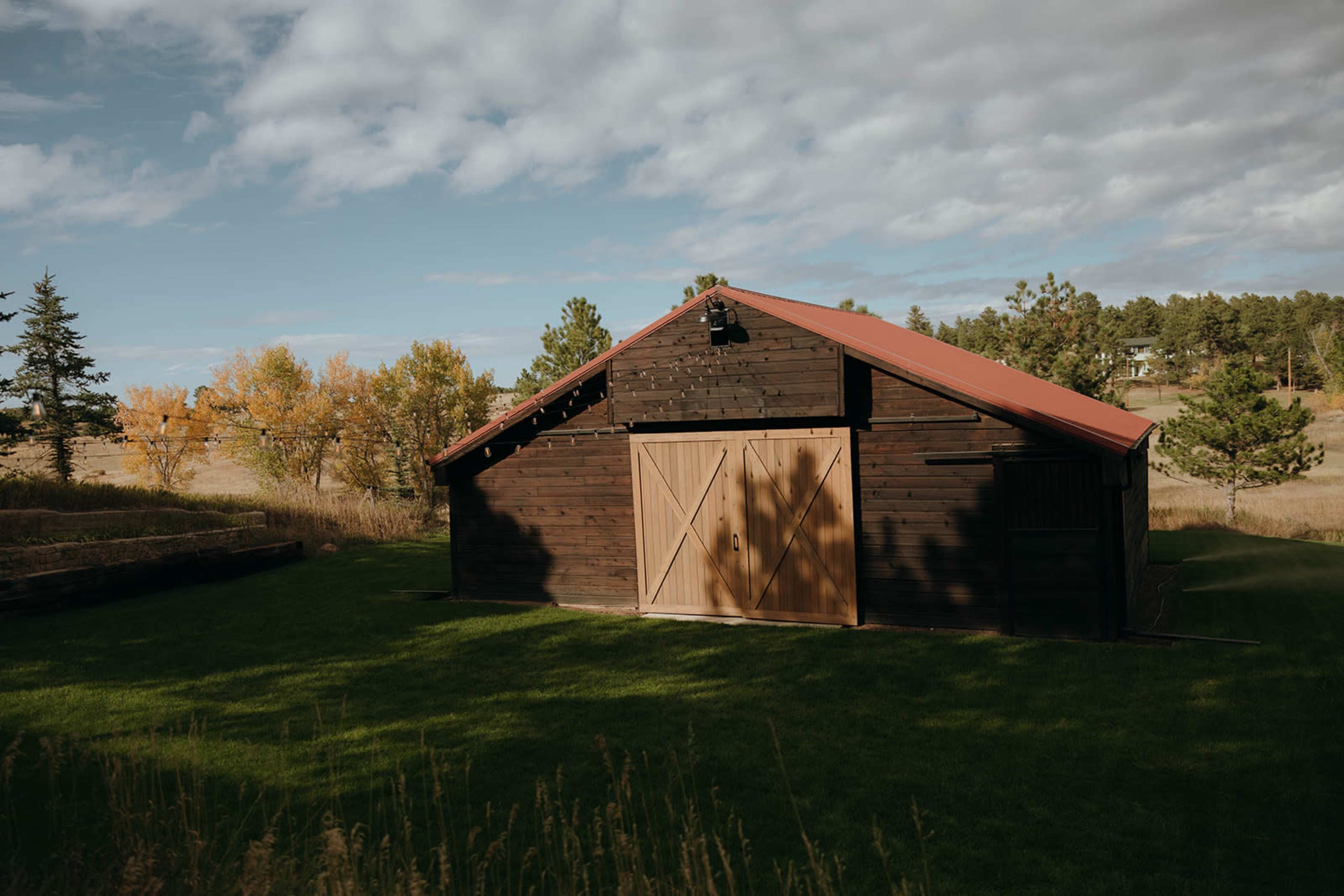 A dark wooden barn with a red roof stands in a grassy area surrounded by trees and open fields.