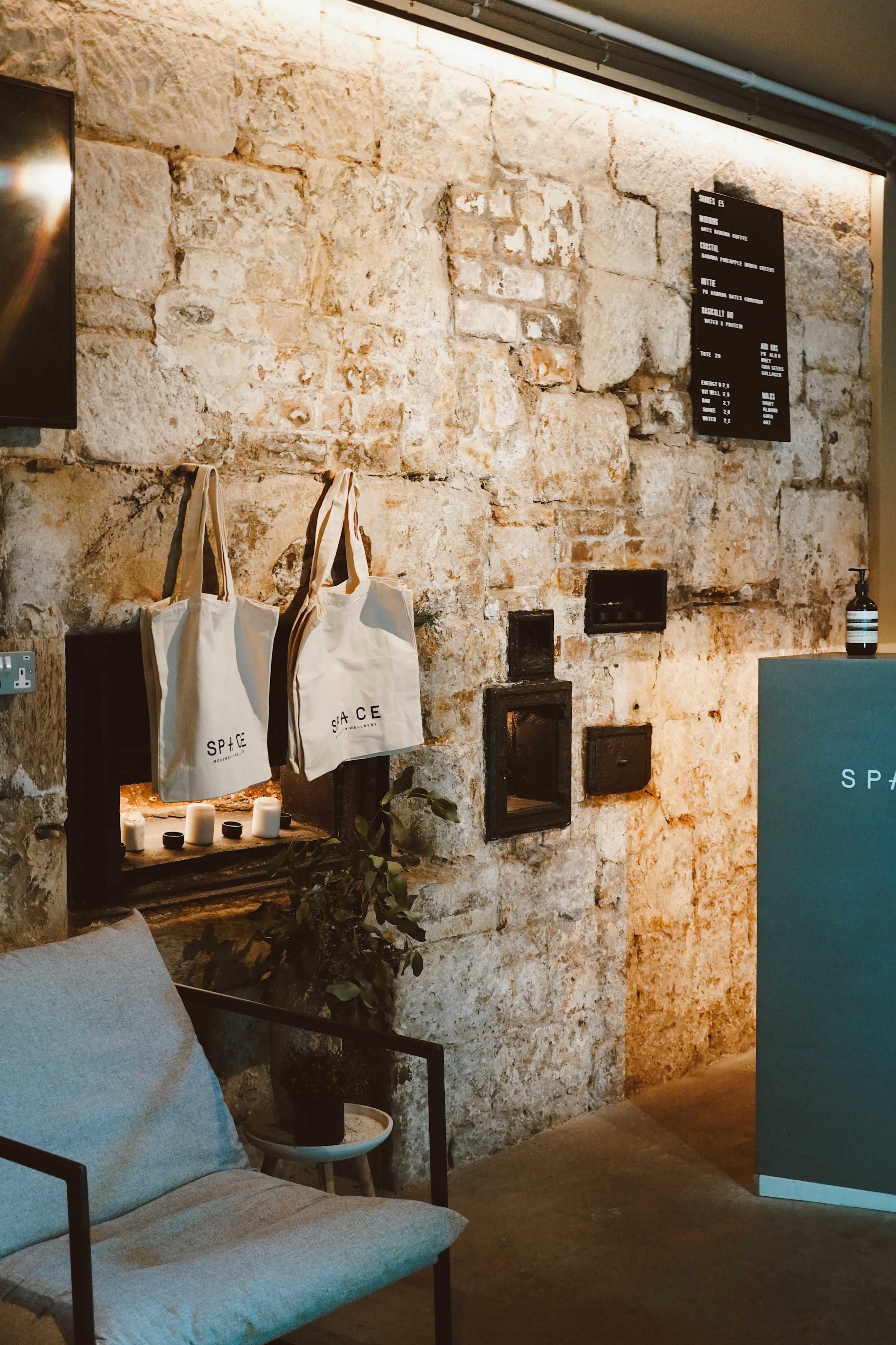 The image shows a cozy interior with a stone wall, a light-colored chair, and tote bags hanging on a hook, along with a menu board displayed above.