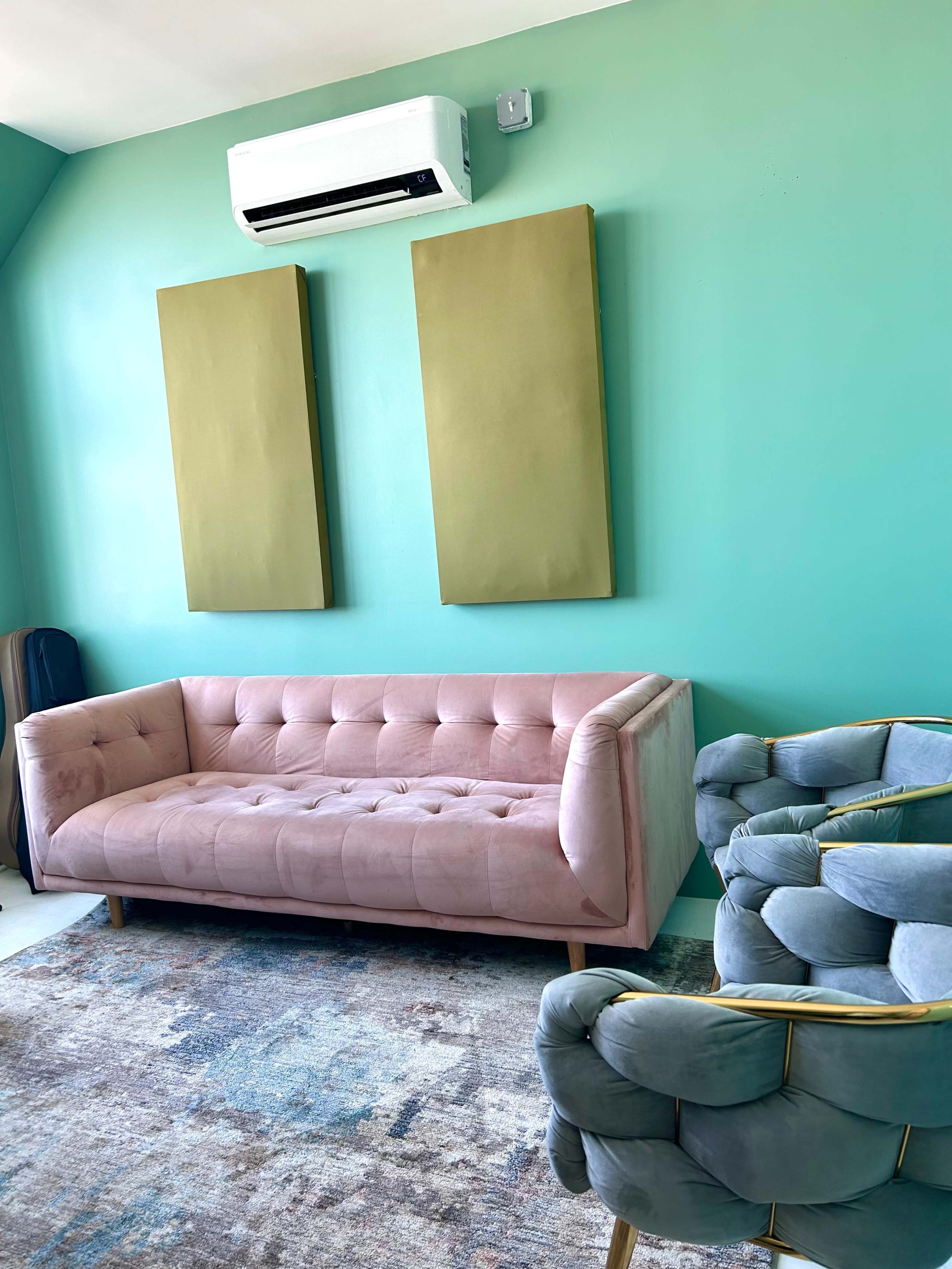 The image shows a brightly colored room featuring a pink tufted sofa, a blue chair, two large gold wall canvases, and an air conditioning unit on the wall.