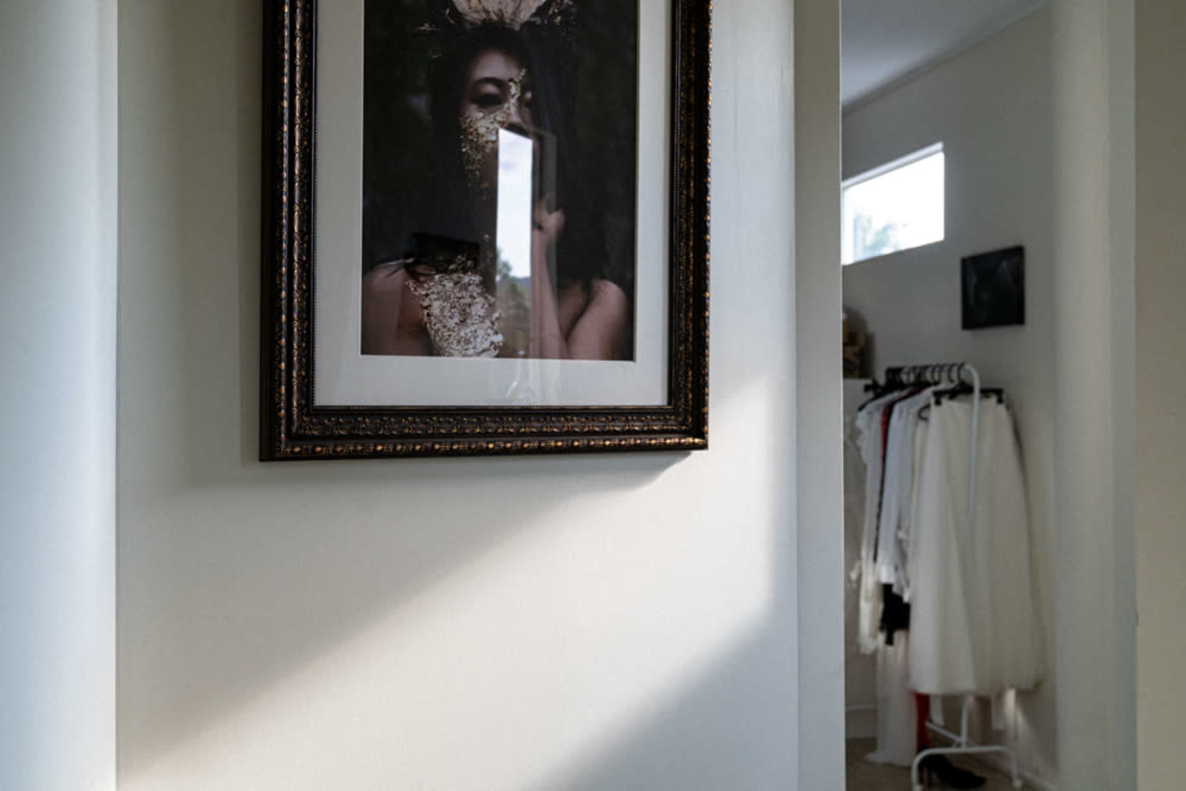 A framed portrait hangs on a white wall, reflecting light from a nearby window, with clothing displayed on a rack in the background.