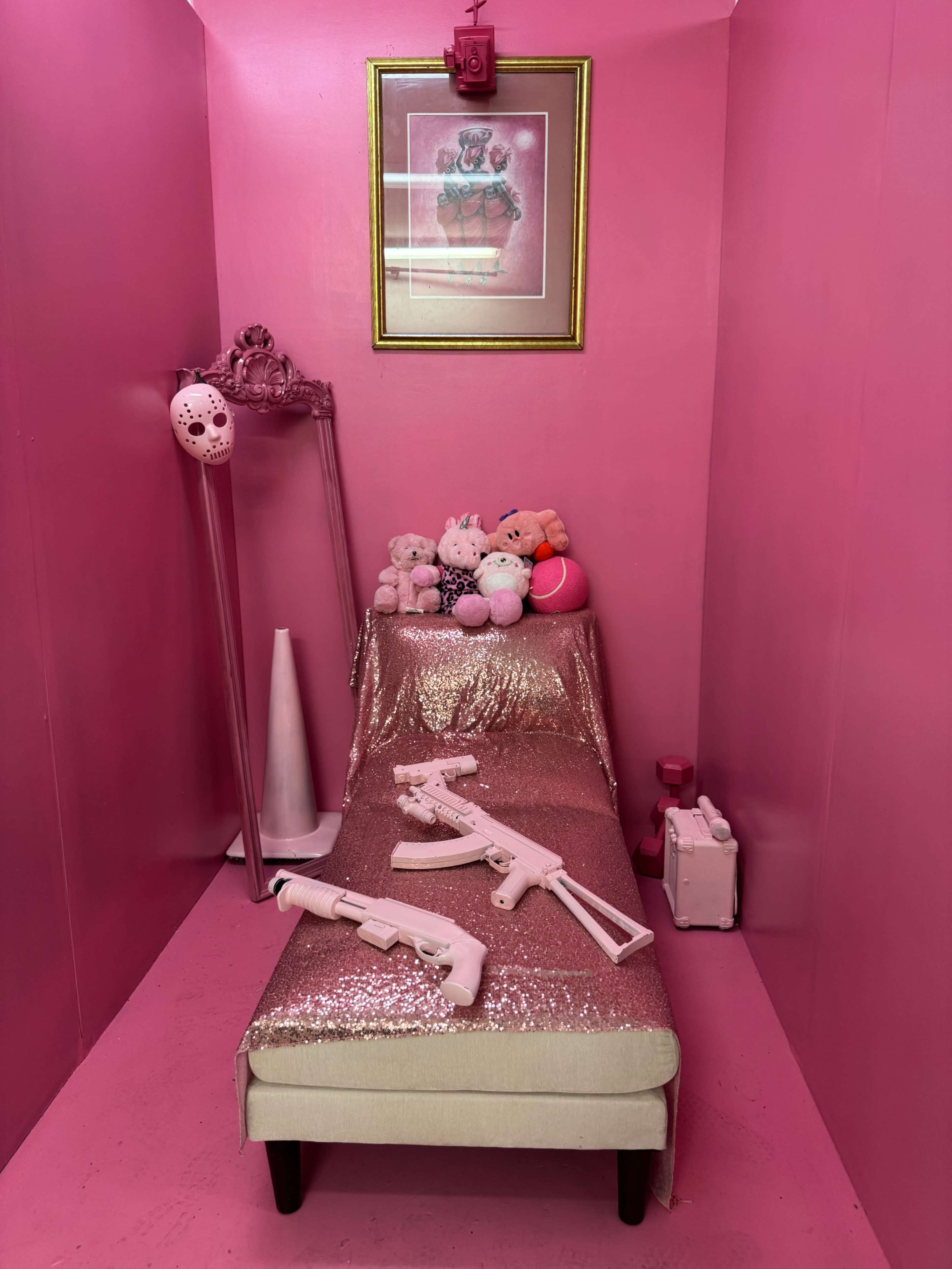 The image shows a small, pink room furnished with a bed covered in a shiny pink fabric, adorned with stuffed animals and two white toy guns.