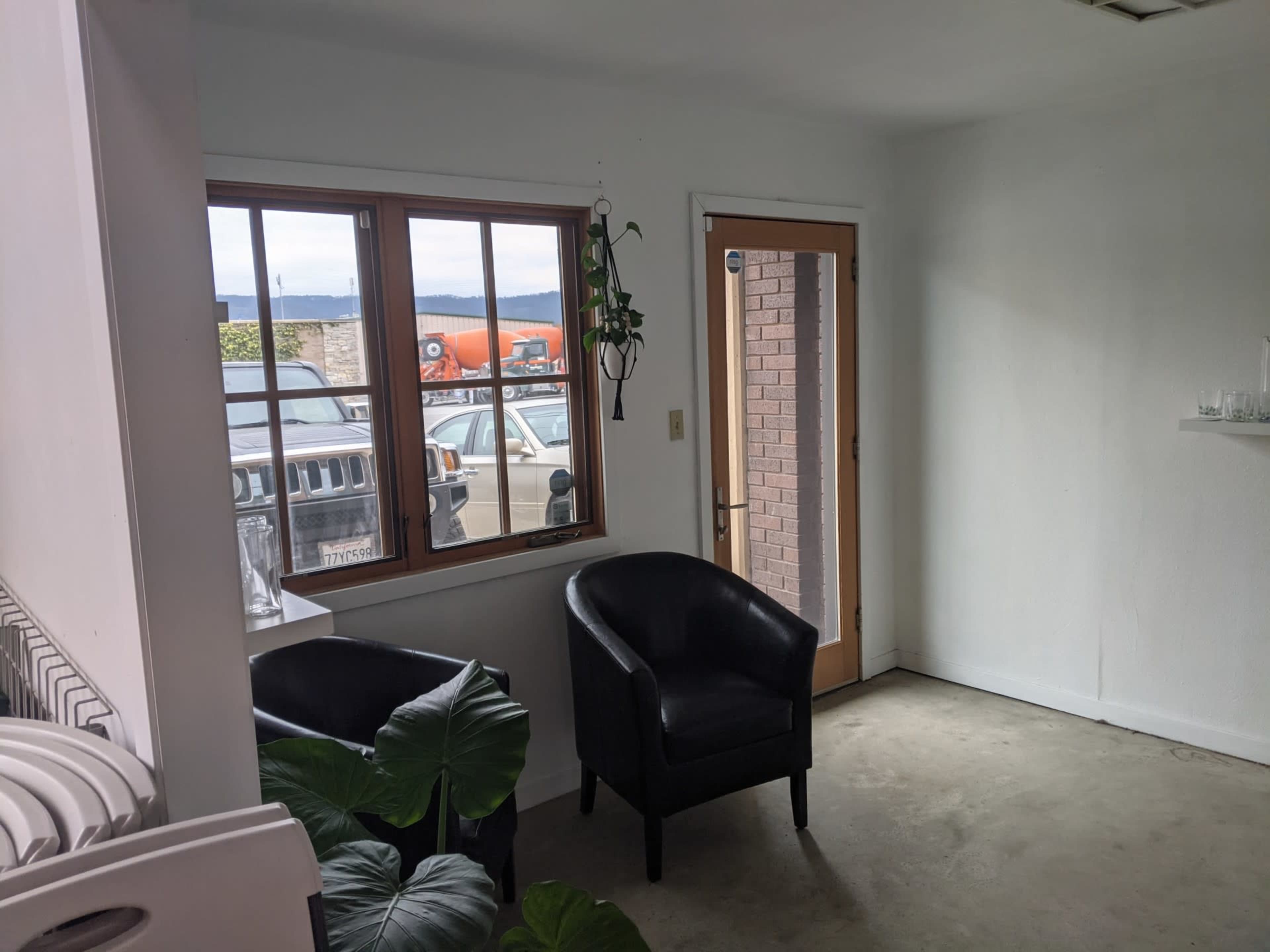A small room with two black chairs, a window revealing parked vehicles outside, and a door leading to another space.