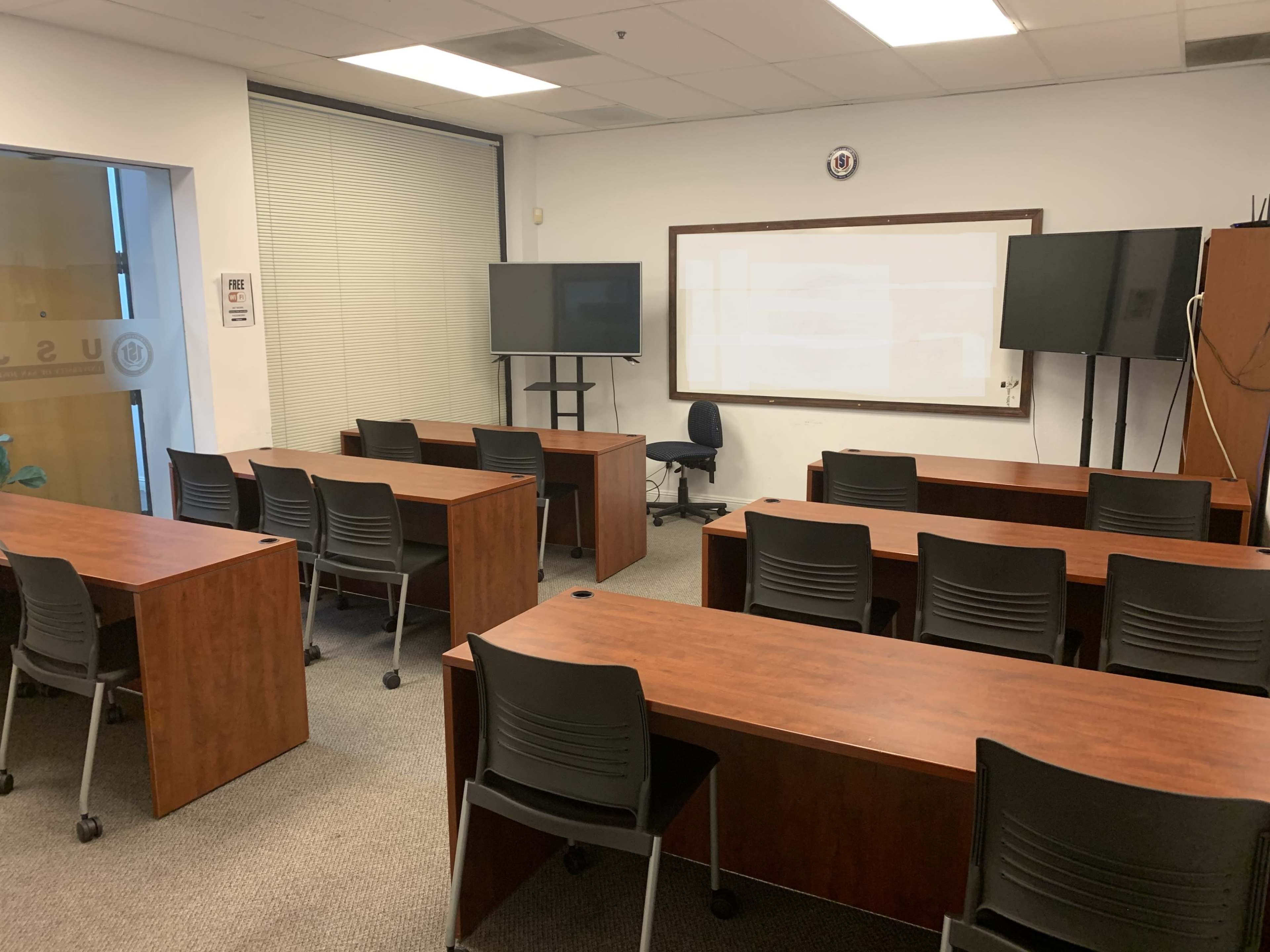 A classroom with rows of wooden tables and black chairs, two monitors positioned at the front, and a whiteboard on the wall.