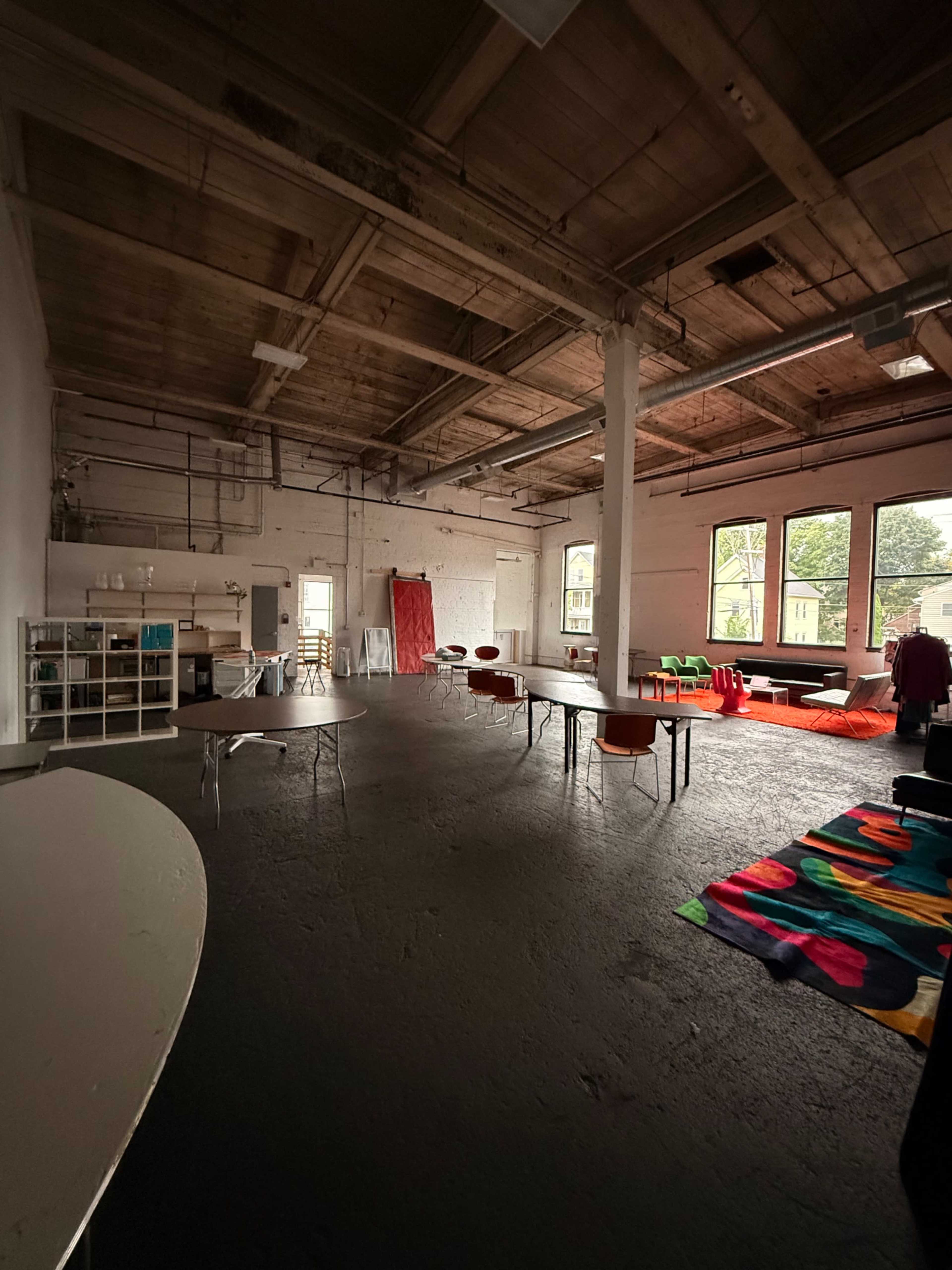 The image depicts a spacious, industrial-style room with large windows, multiple tables, and colorful areas on the floor.