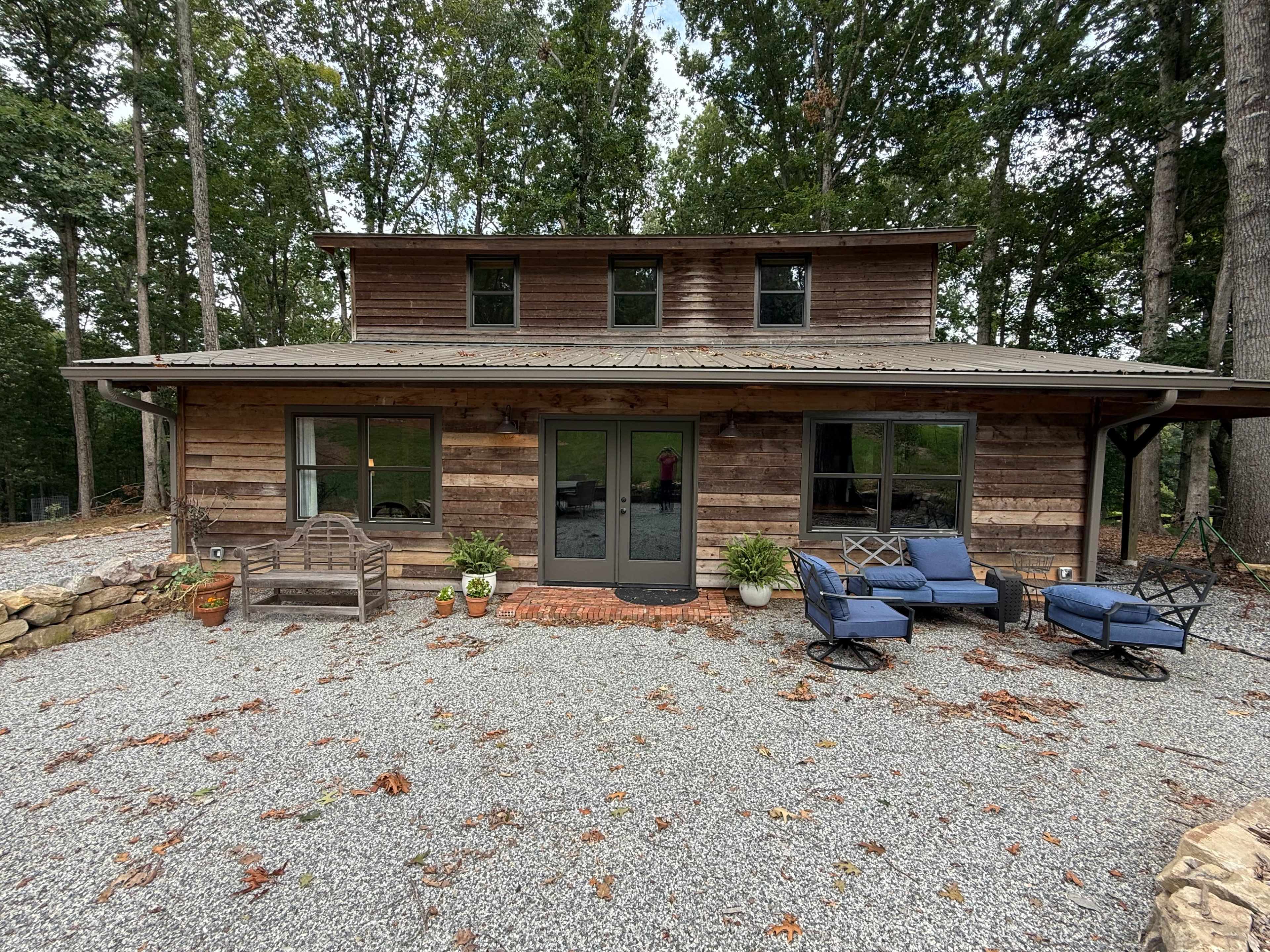 A wooden cabin with a gravel patio is surrounded by trees, featuring large windows and a front entrance with seating.