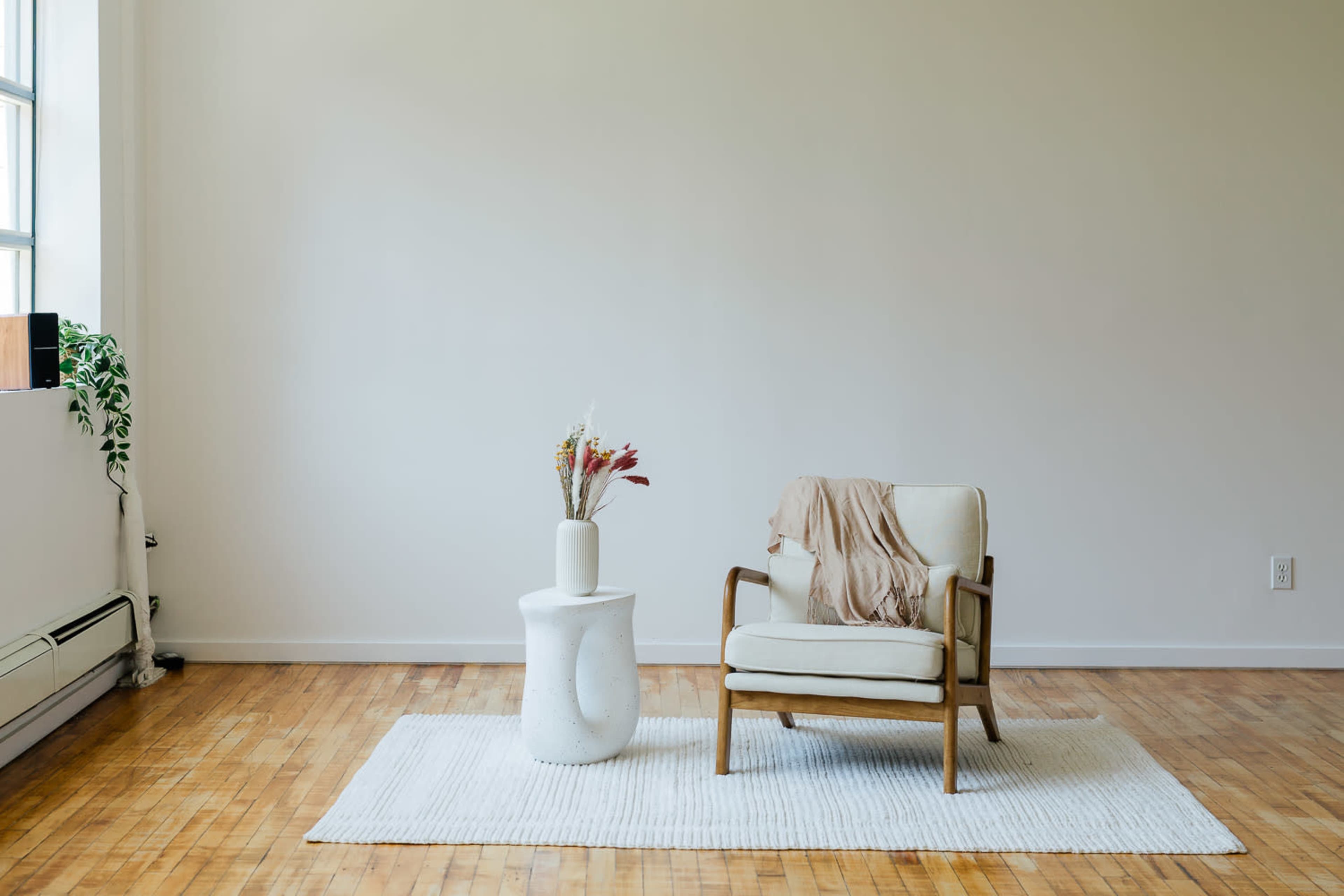 The image features a minimalist living space with a wooden chair, a small round table, and a decorative vase atop a white area rug.
