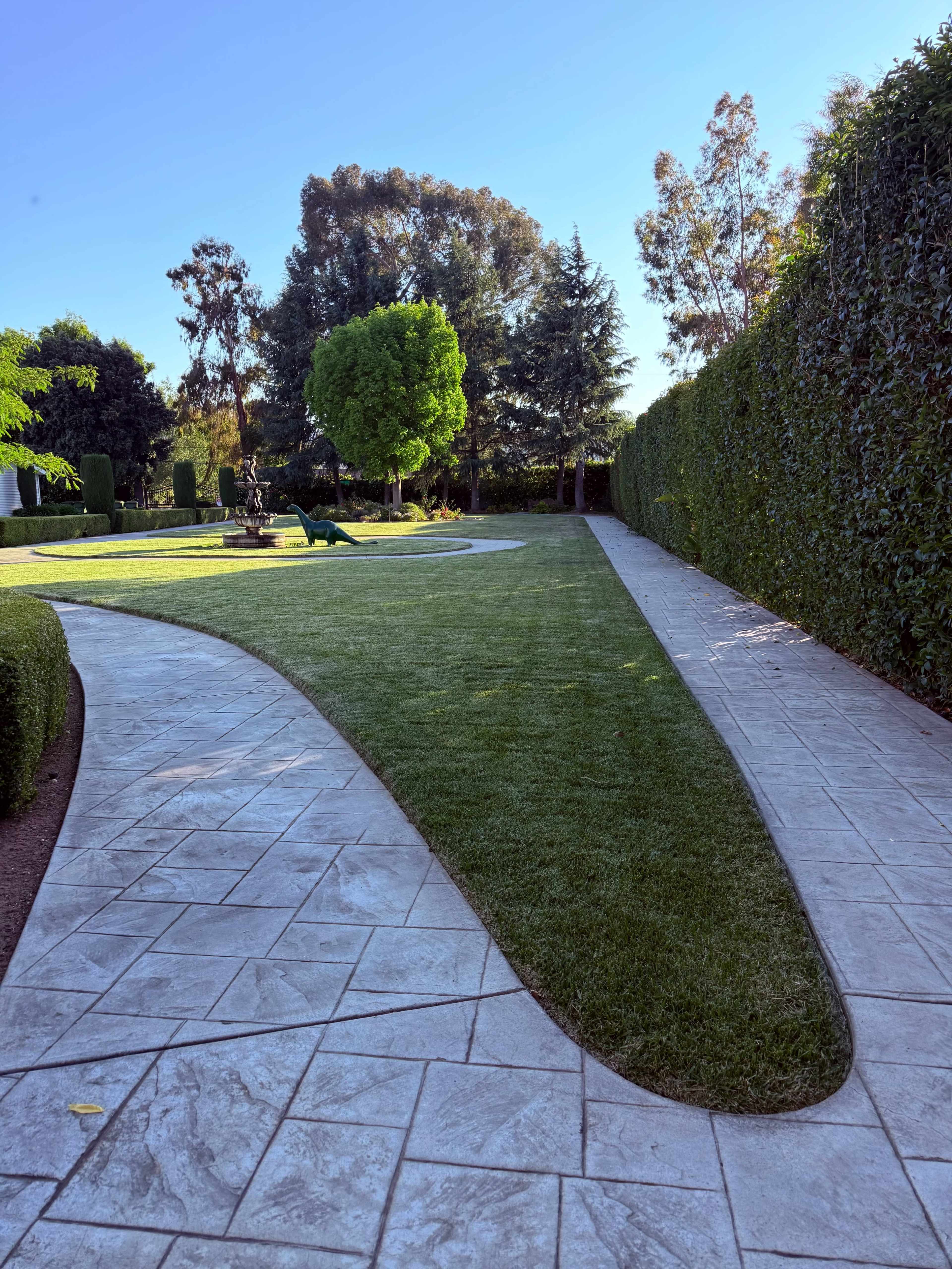 The image shows a manicured lawn bordered by a curved concrete pathway, surrounded by neatly trimmed hedges and trees.