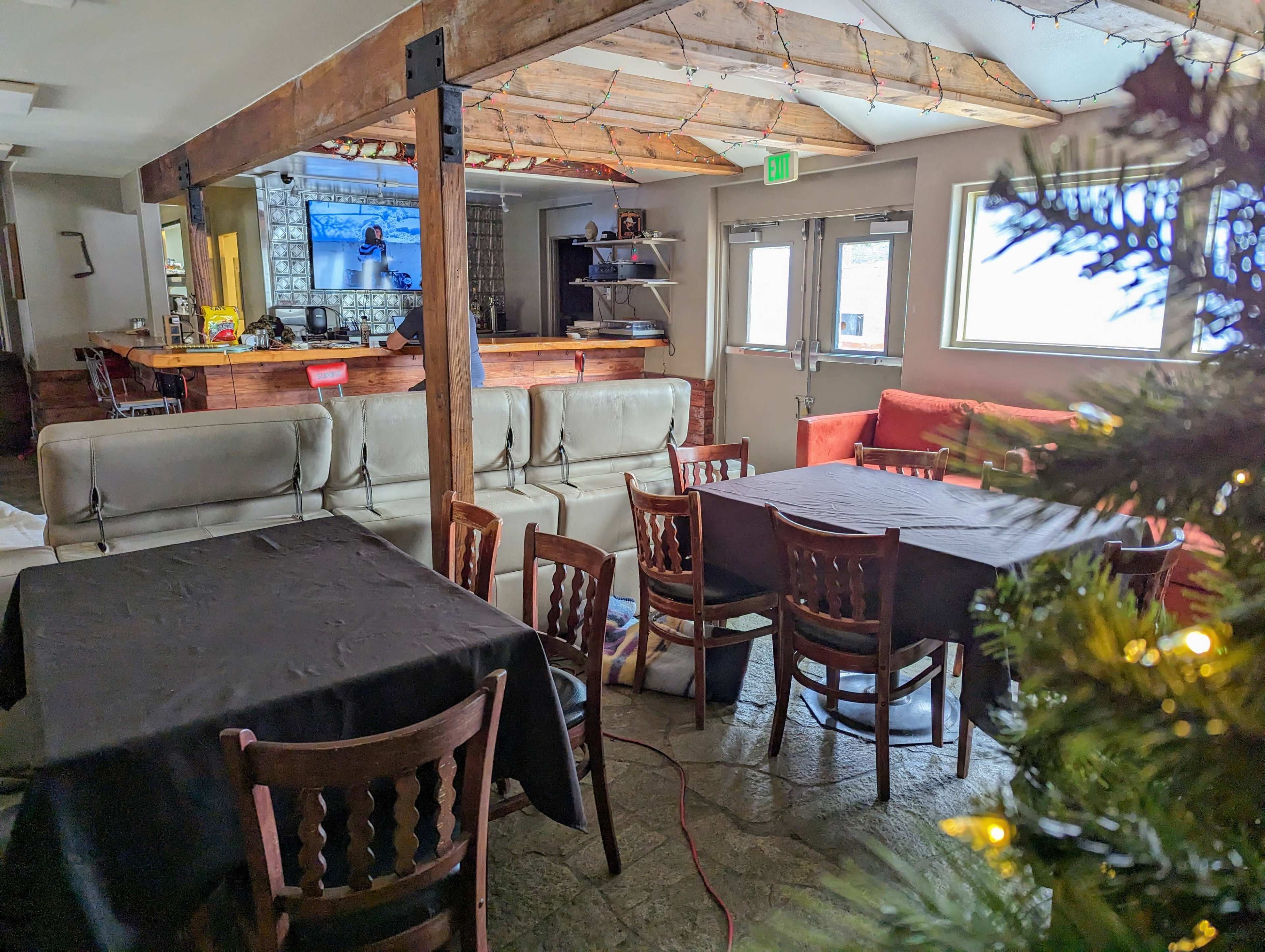 The image shows a cozy interior space with several tables and chairs, a sofa, and a visible television in the background.