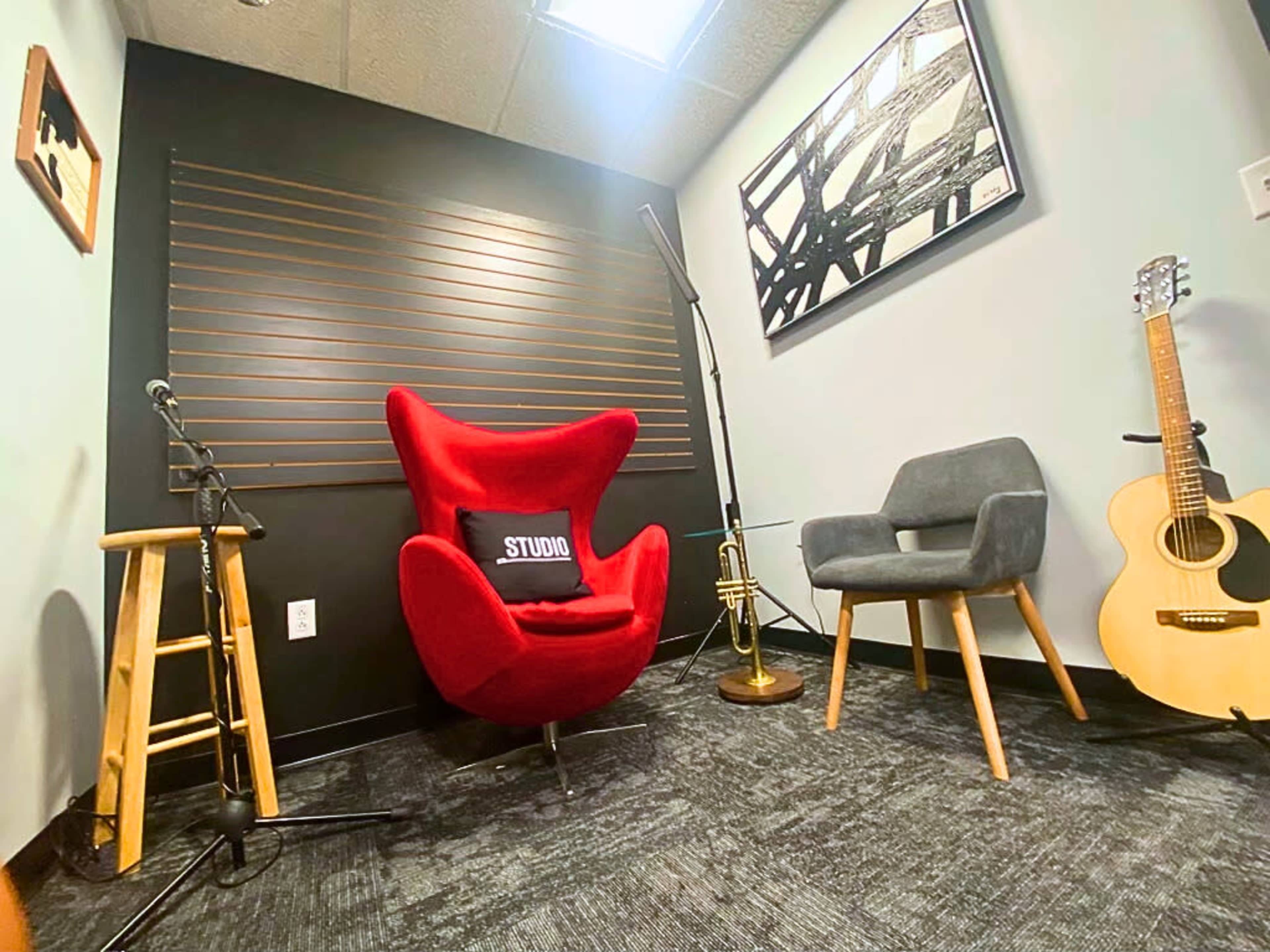 A cozy recording studio corner featuring a red chair, a gray chair, a guitar, a trumpet, a microphone stand, and artwork on the wall.