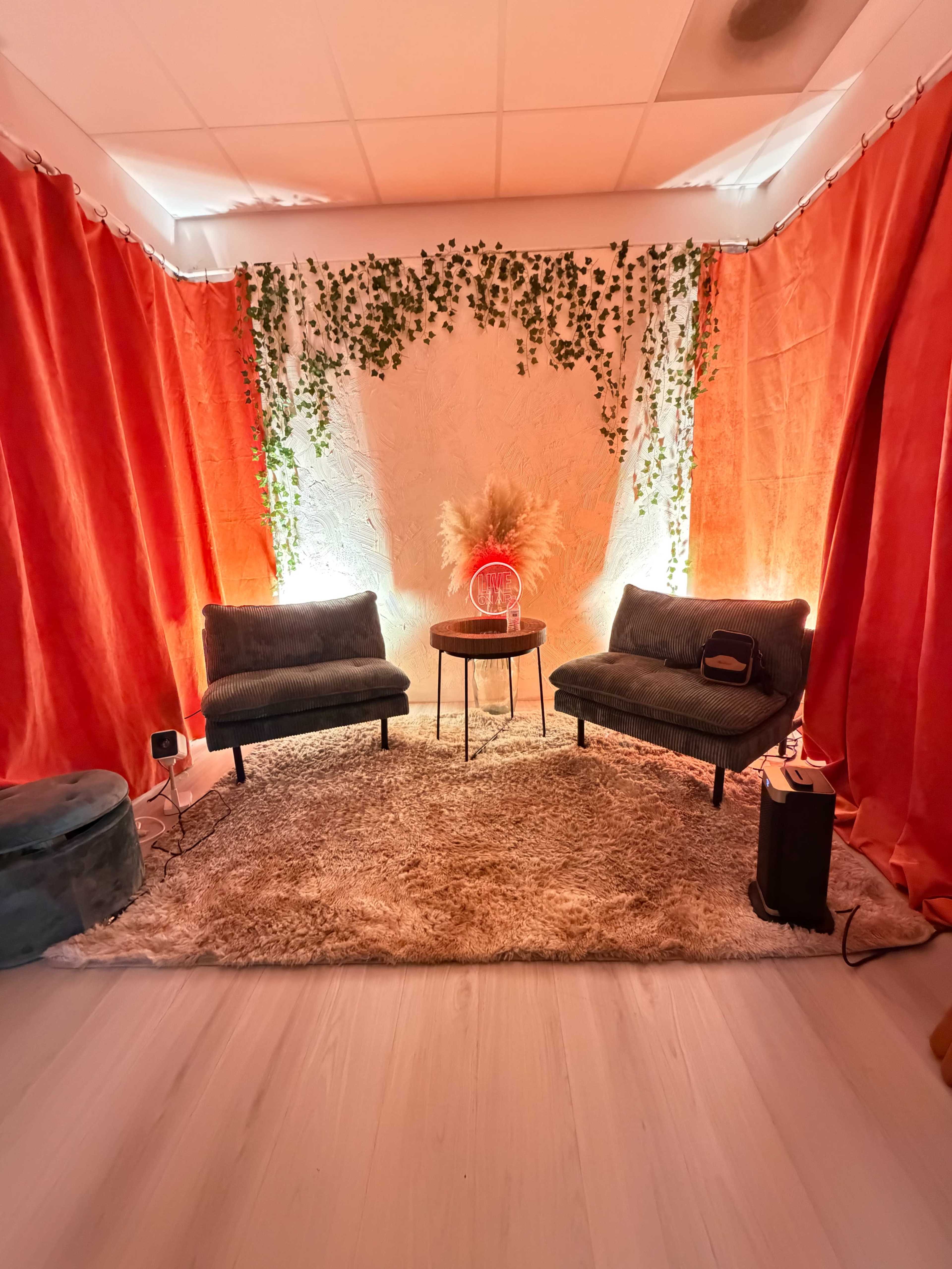 The image shows a cozy living space with two dark grey sofas, a round table, and decorative elements including a textured rug, lighting, and artificial plants against bright orange curtains.