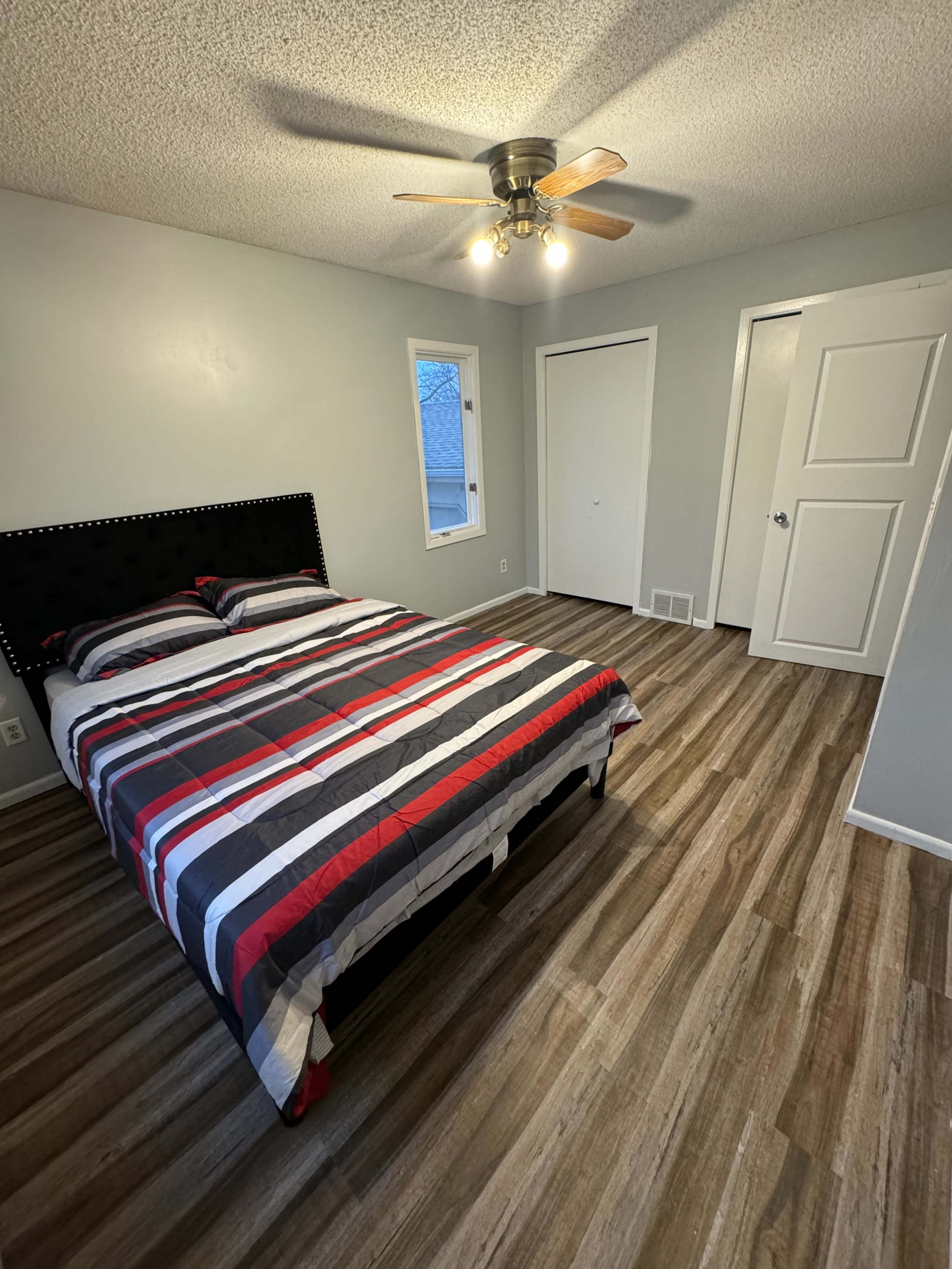 A bedroom features a striped bedspread on a bed with a black headboard, wooden flooring, and two windows.