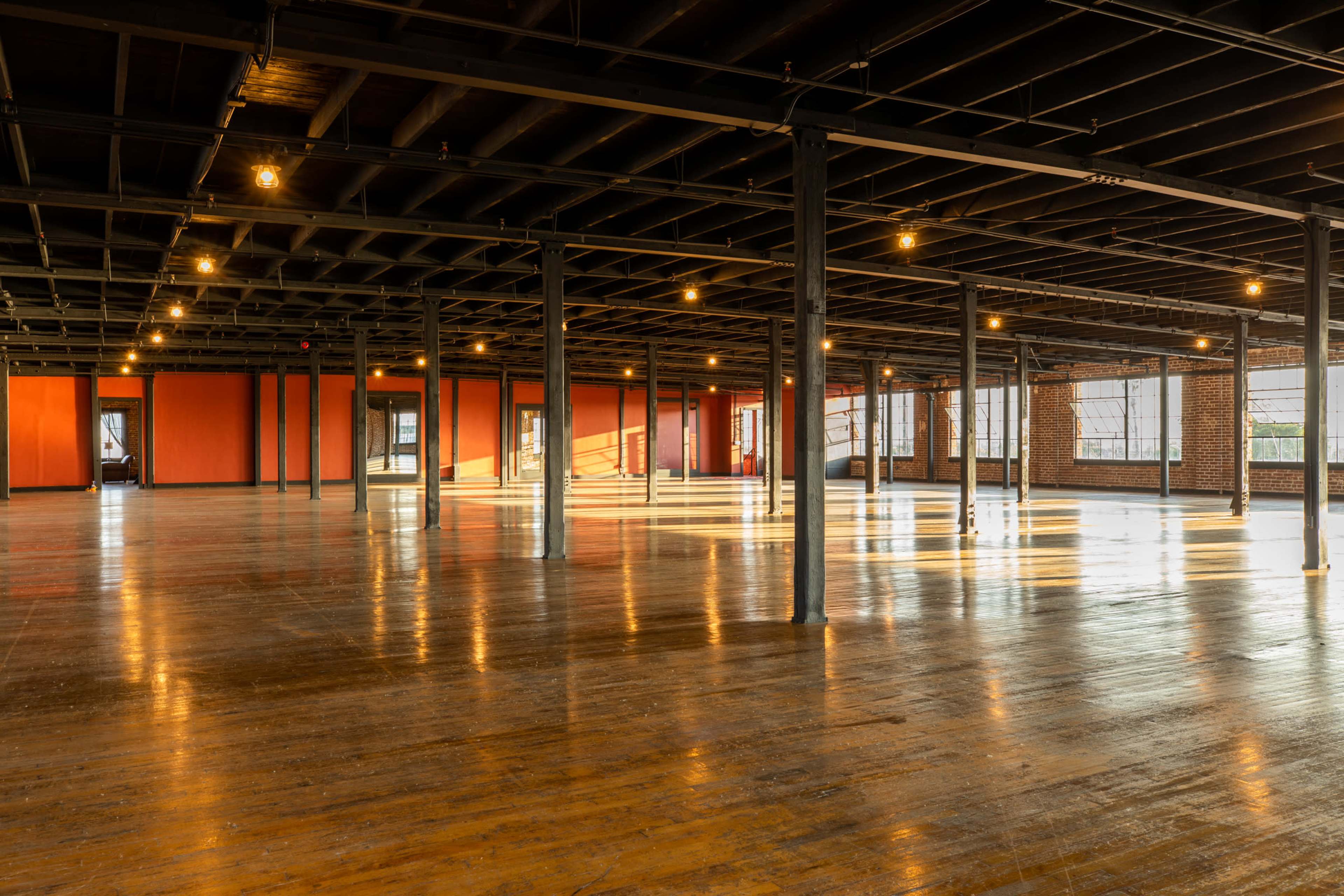 A spacious, empty industrial-style room with wooden floors, exposed beams, and large windows allowing natural light to fill the space.