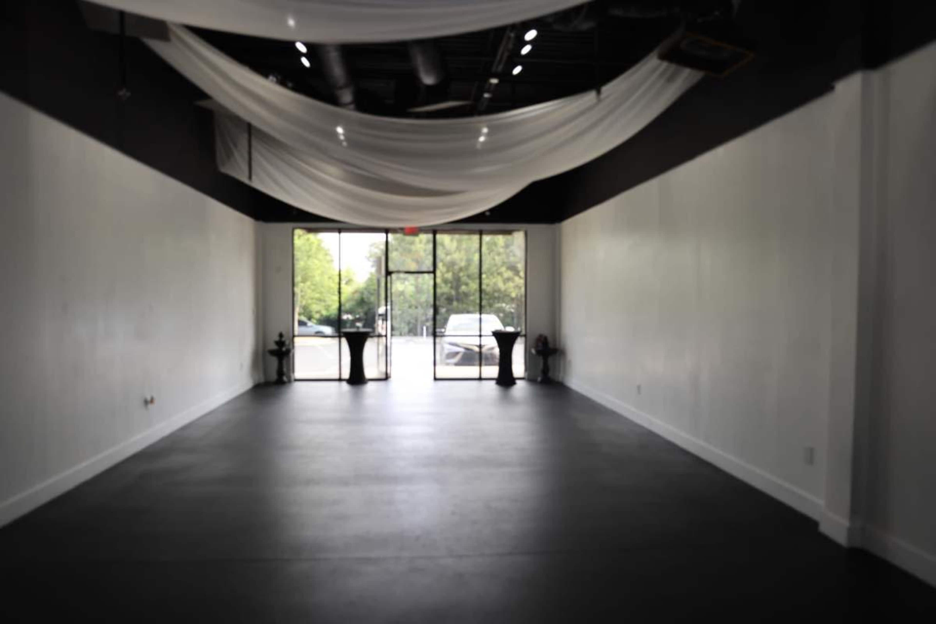 A spacious, empty room with a black floor, large front windows, and draped fabric overhead.