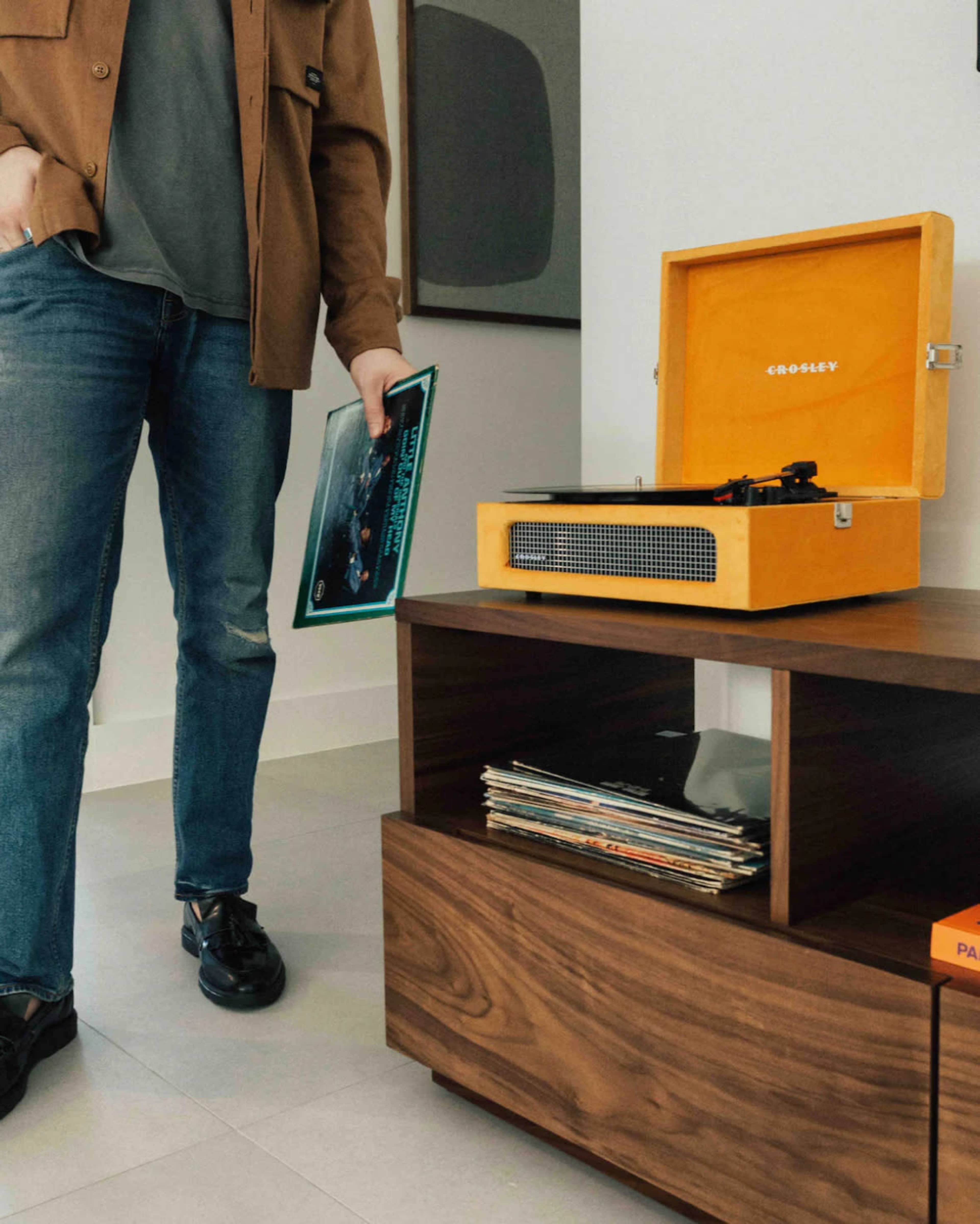 A person stands next to a wooden record player table, holding an album cover while a yellow turntable is open on the surface.
