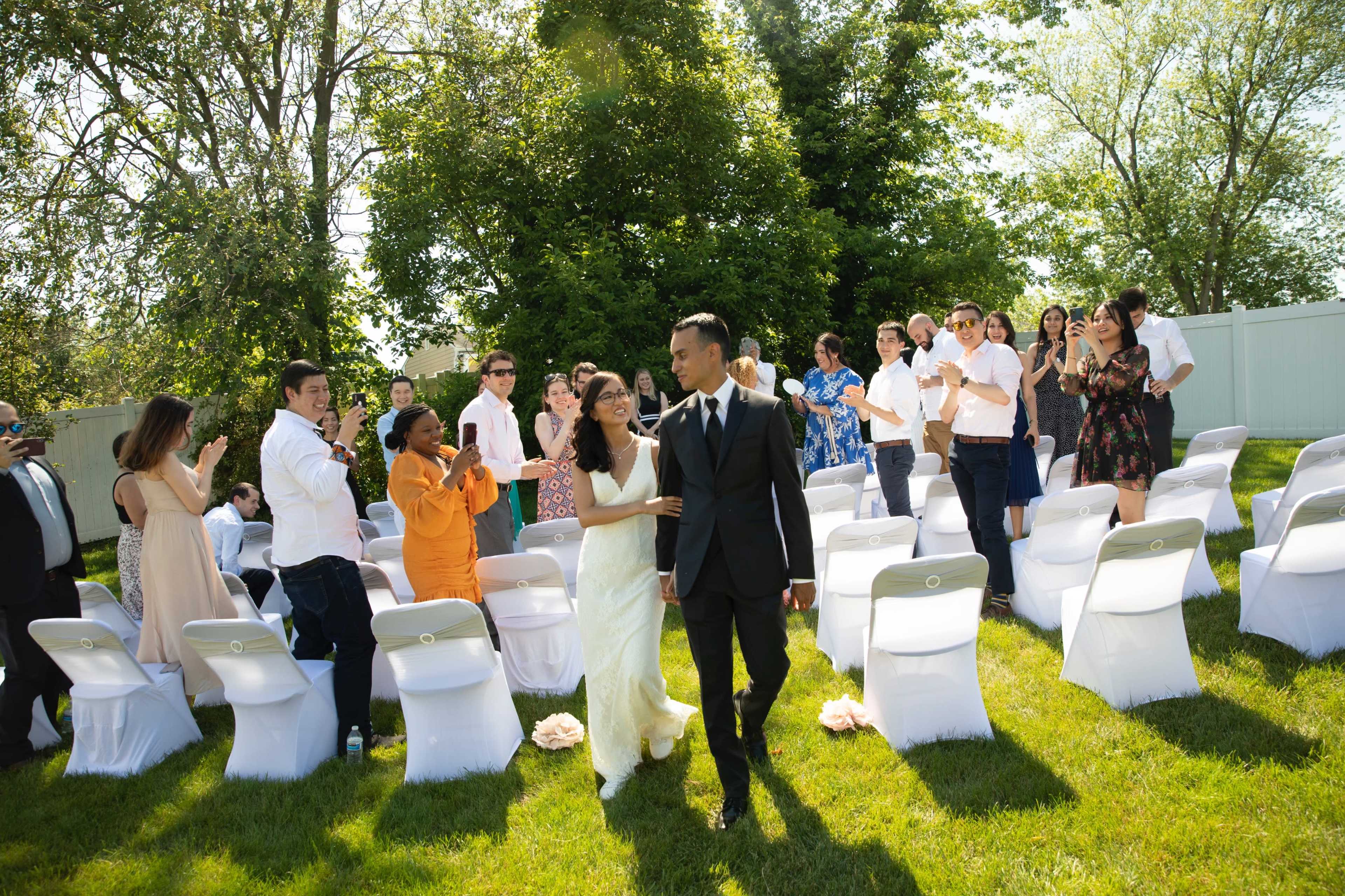 A bride and groom walk down the aisle surrounded by seated guests in a sunny outdoor wedding ceremony.