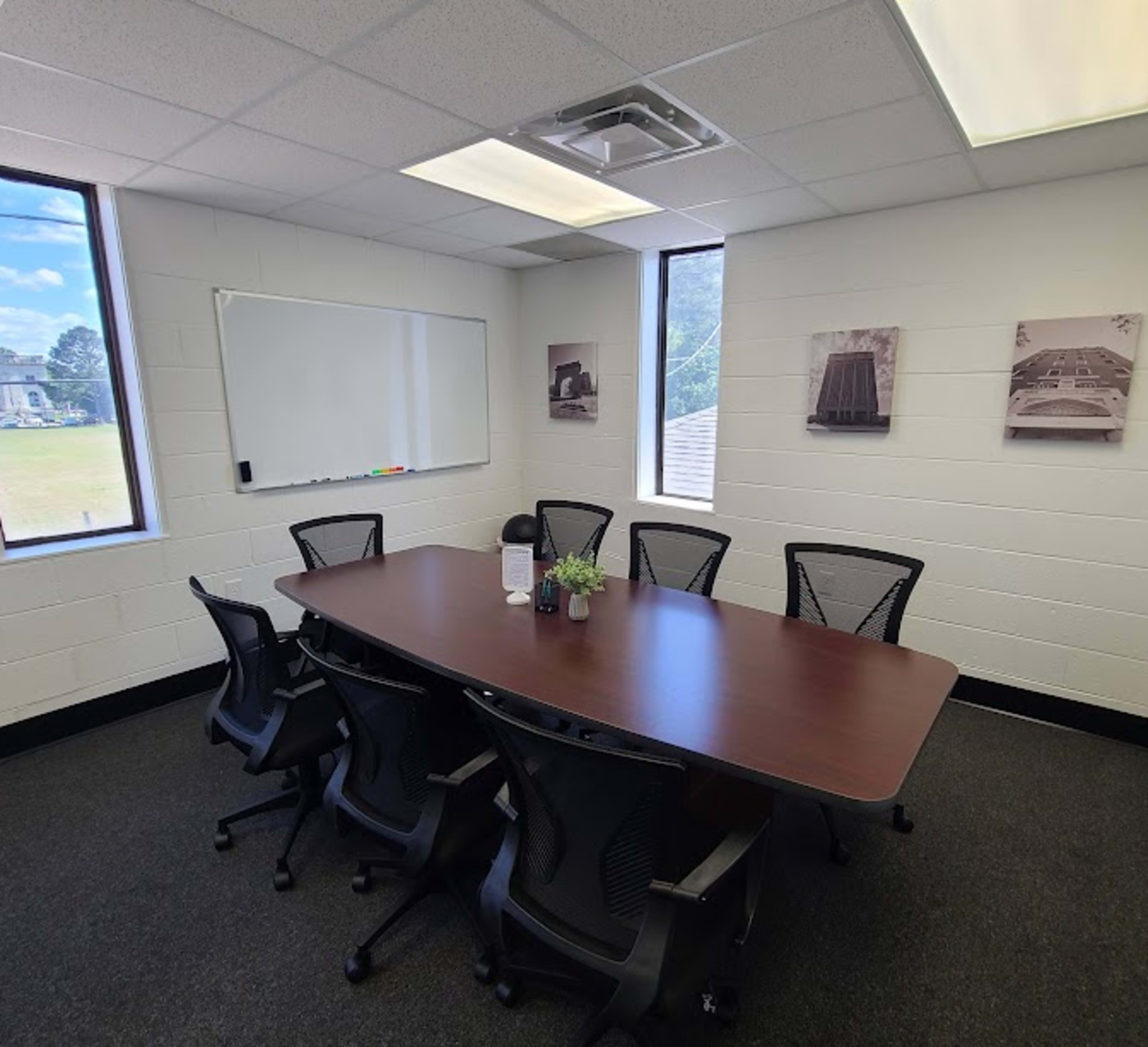 The image shows a conference room with a large wooden table surrounded by six black office chairs, a whiteboard, and framed pictures on the walls.