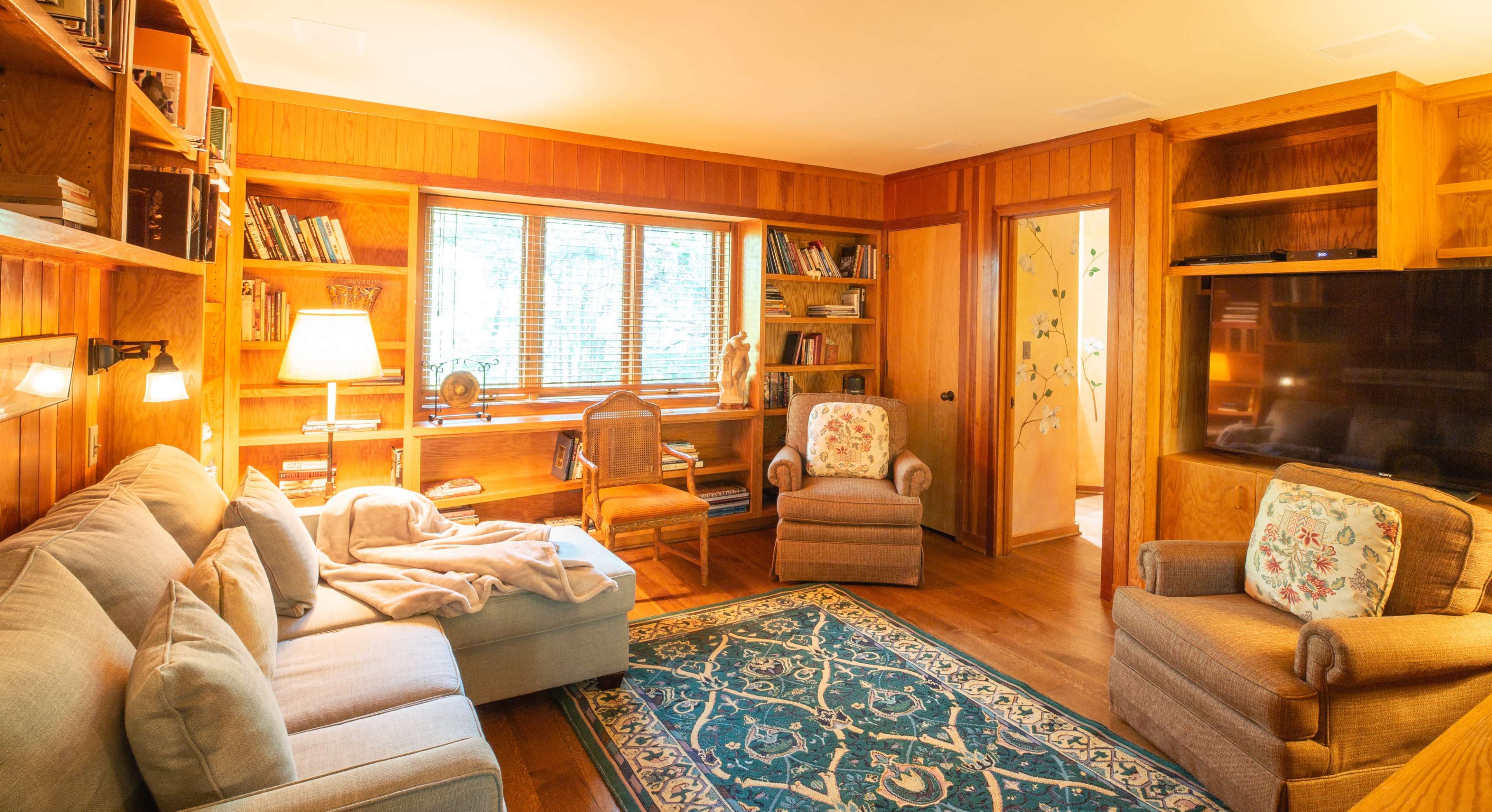 A cozy living room with a sofa and two armchairs, surrounded by wooden bookshelves and a television.