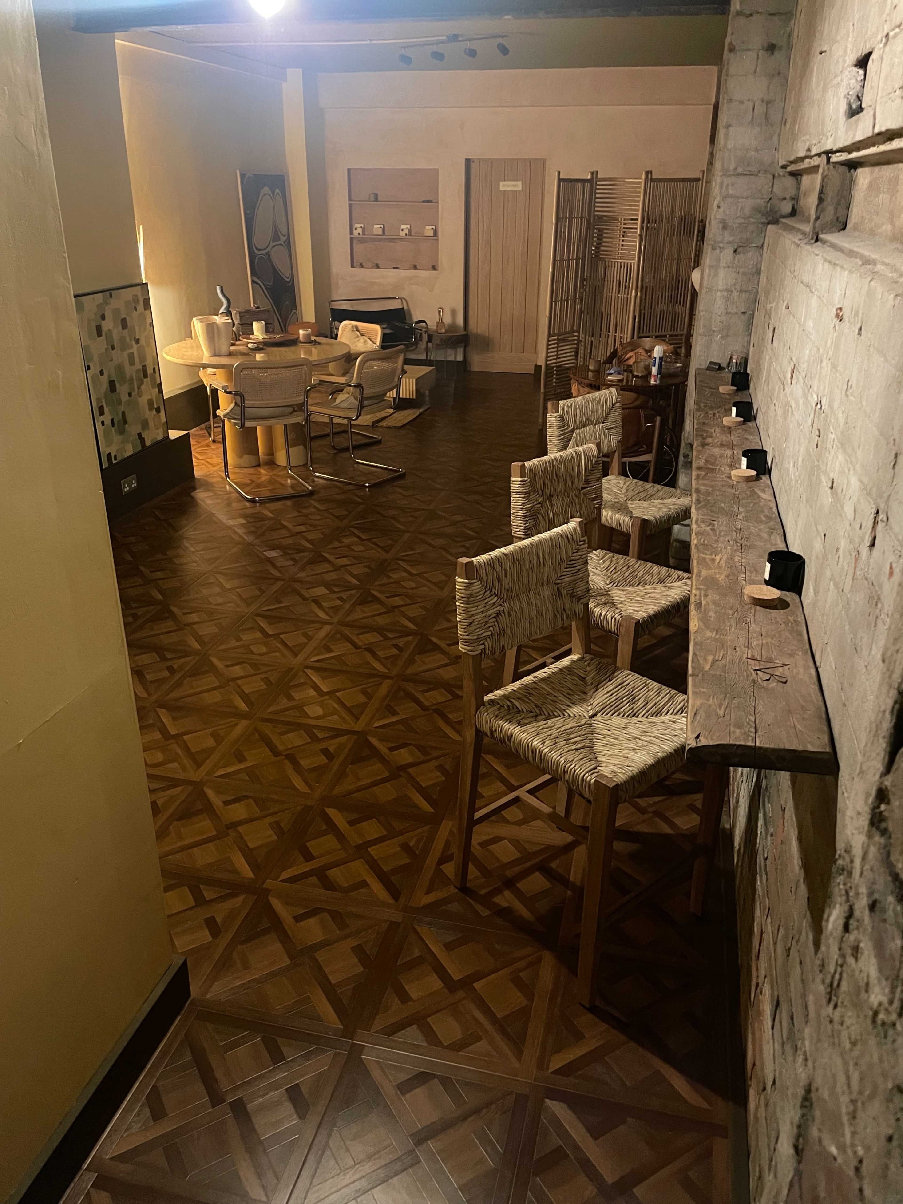 A spacious interior features a wooden floor with a geometric pattern, several chairs around a round table, and a long counter with bar stools against a wall.