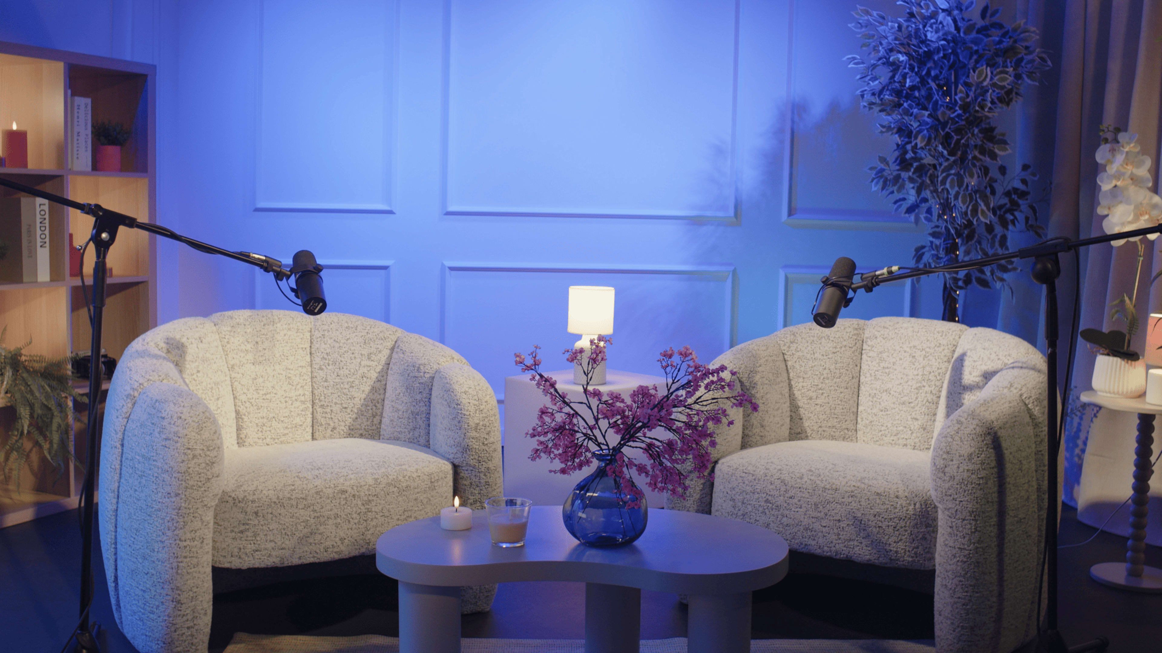 The scene features two circular armchairs facing each other on a stage, with a small table between them holding a candle and a vase of flowers, all set against a softly lit blue background.