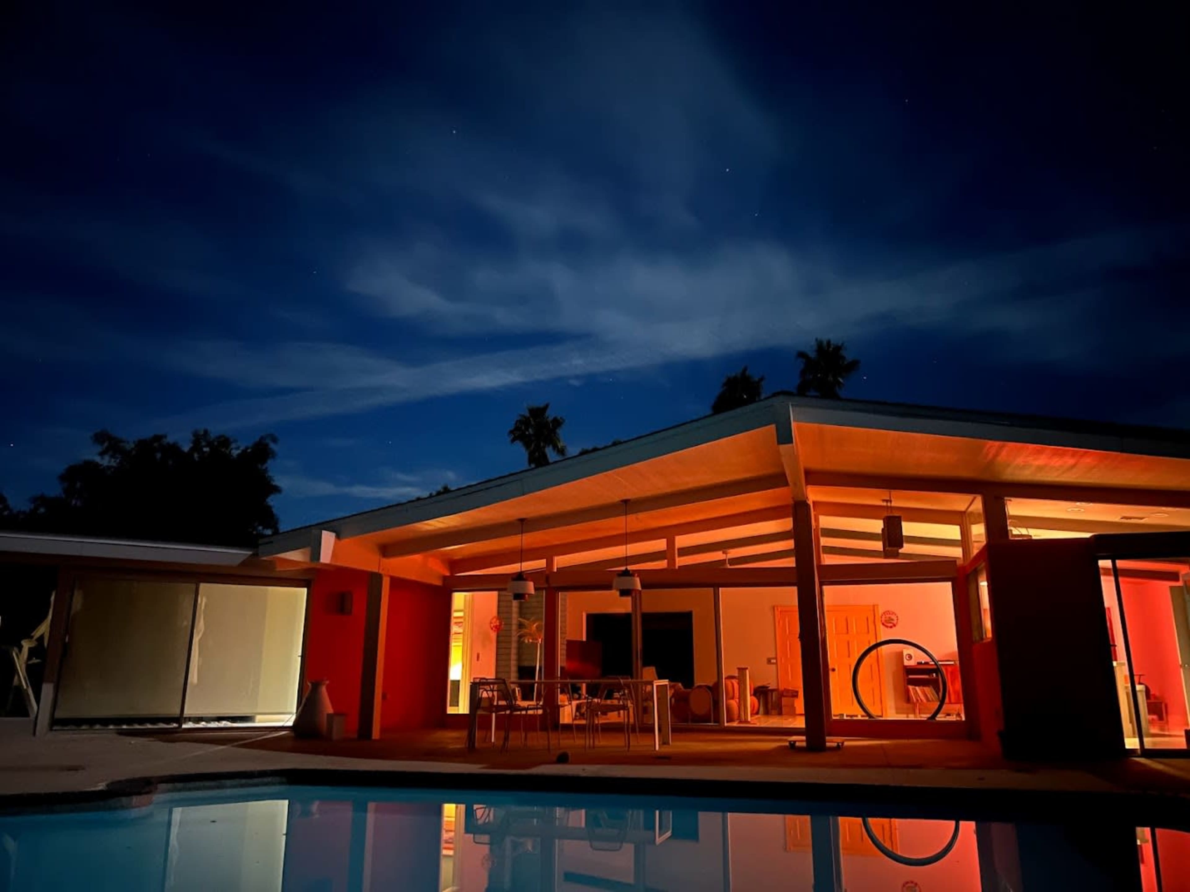 A modern house with large windows is illuminated from within, reflecting in a pool at night under a starry sky.