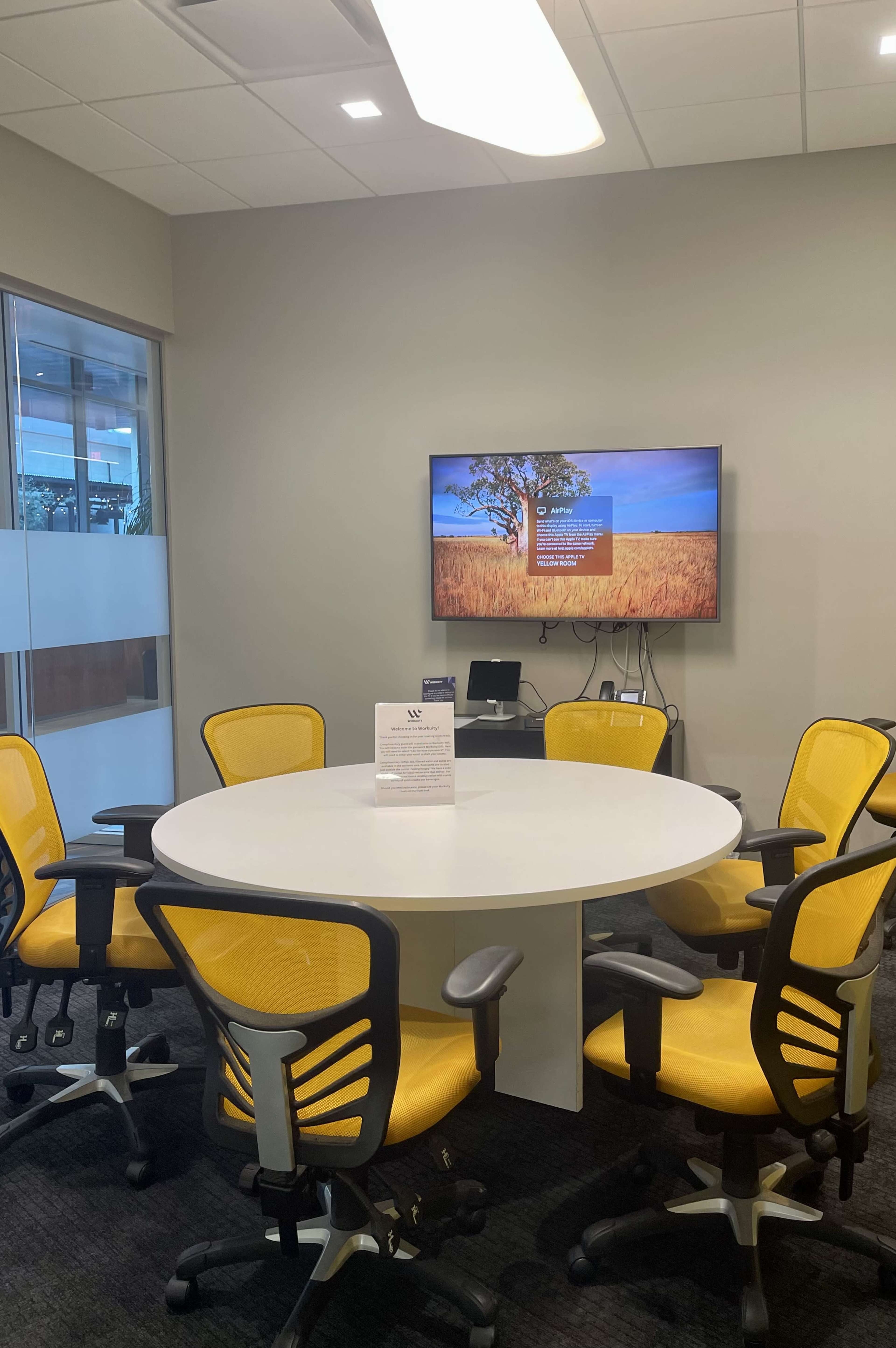 A modern meeting room features a circular white table surrounded by six yellow chairs and a wall-mounted TV displaying a landscape image.