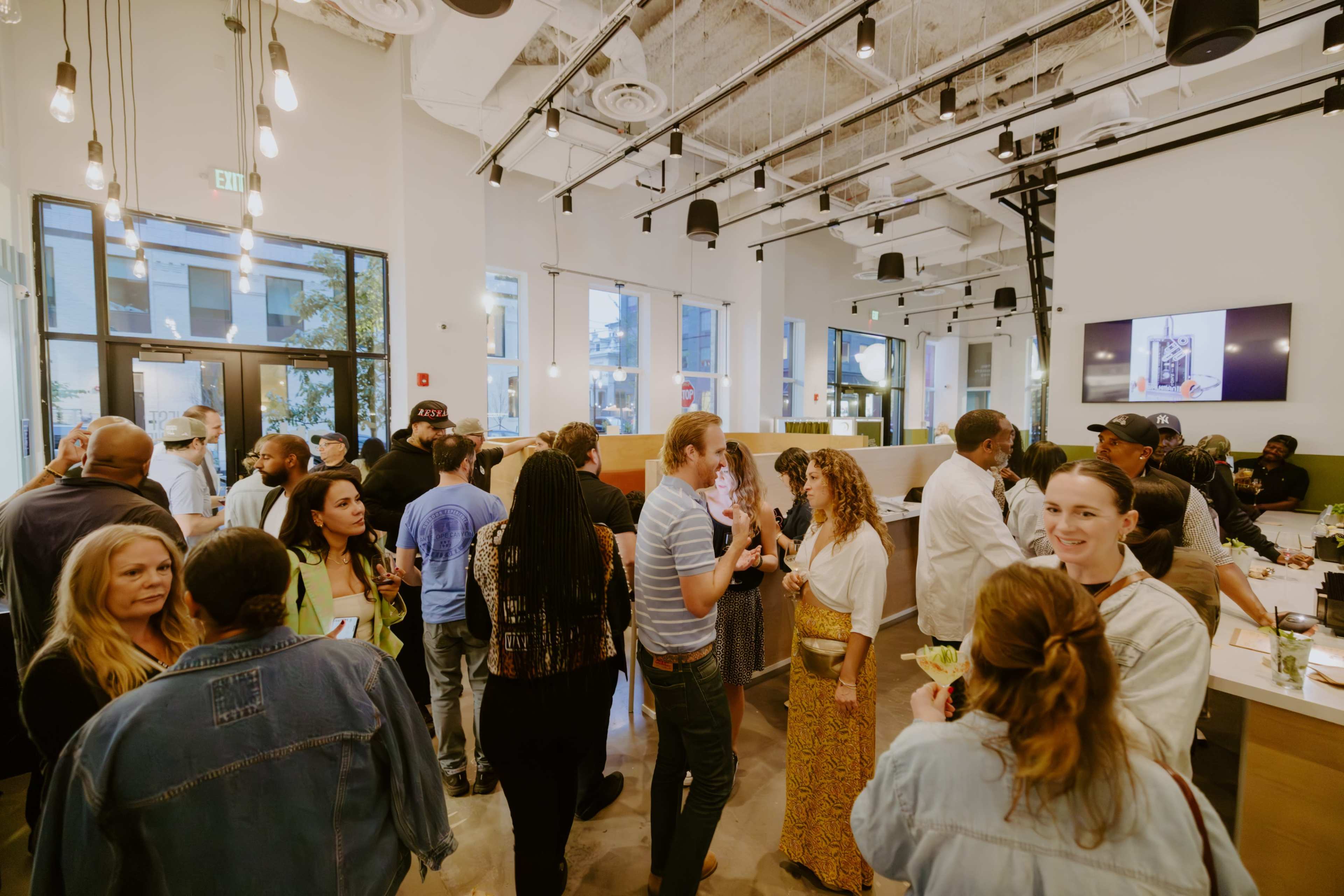 A diverse group of people socialize in a well-lit, modern indoor space with a bar area and large windows.