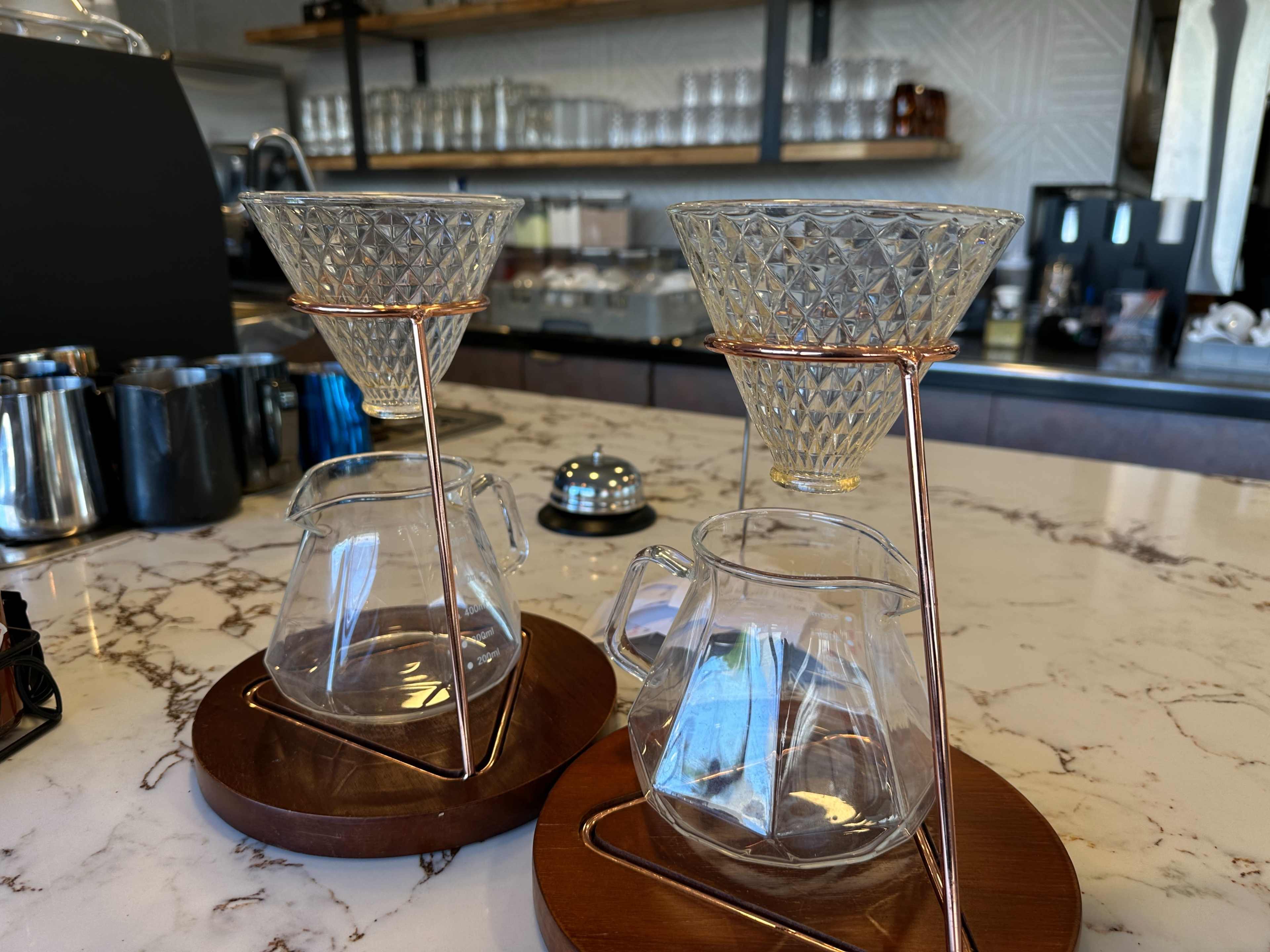 The image shows two glass coffee drippers set atop clear glass carafes on wooden saucers, placed on a marble countertop in a café setting.